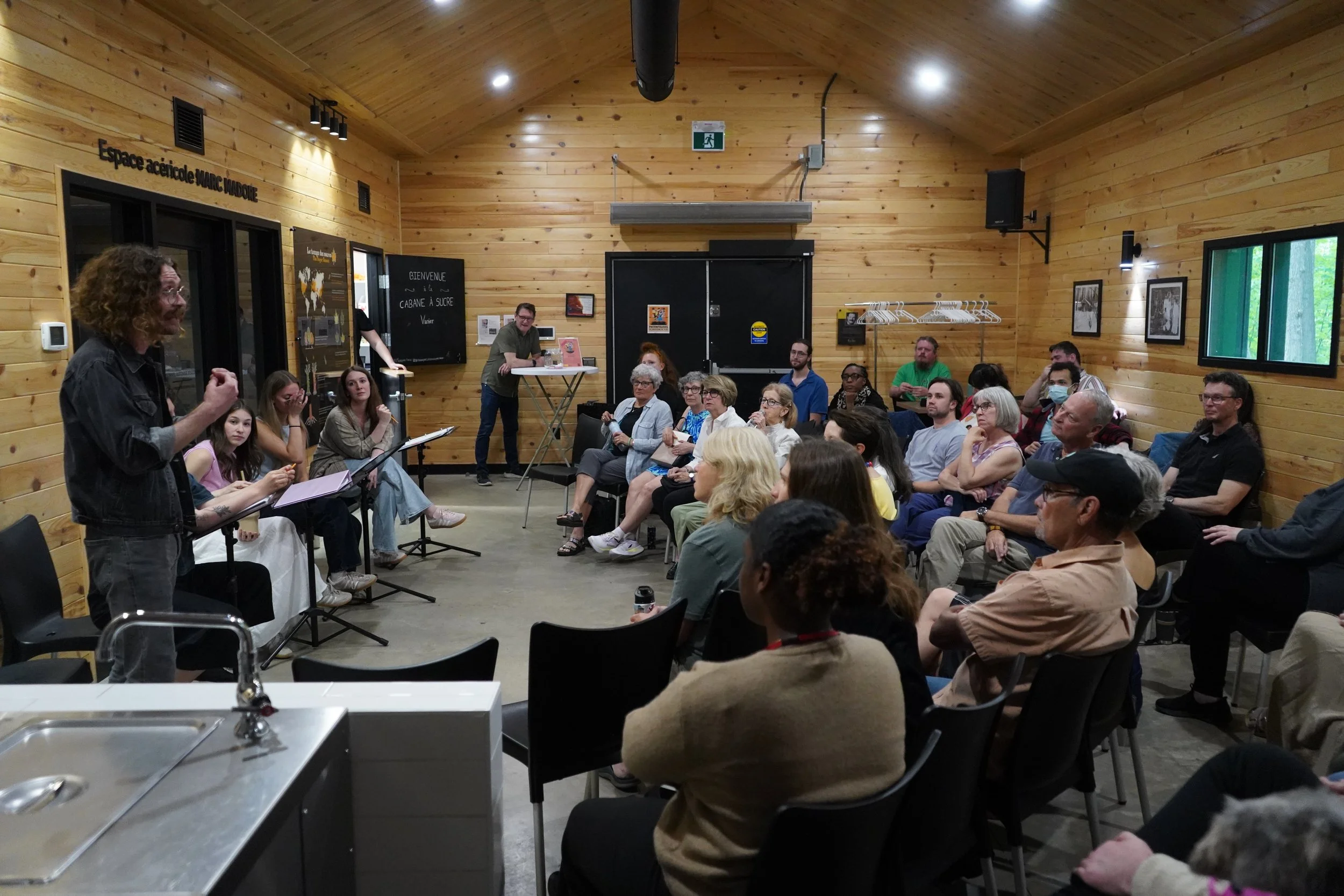 Une réunion ou un atelier se déroule dans une salle en bois, avec des personnes assises en face d'un homme qui semble parler ou présenter. La salle comporte des affiches, un tableau noir, et des fenêtres laissant entrer la lumière naturelle.