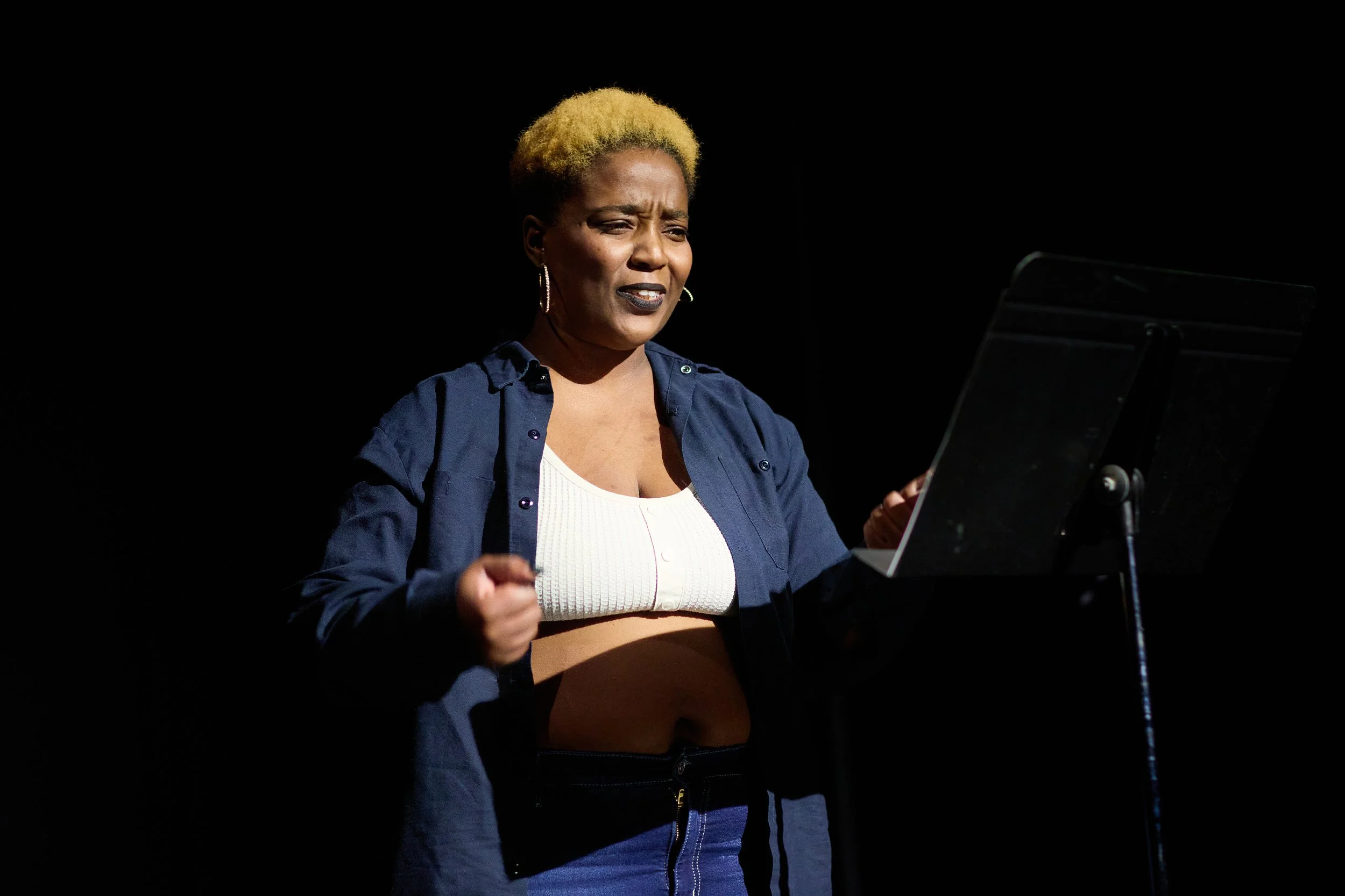 Une femme avec les cheveux courts blonds, portant une chemise bleue, un débardeur blanc et un pantalon noir, semble en train de chanter ou de parler devant un pupitre, avec un fond noir.