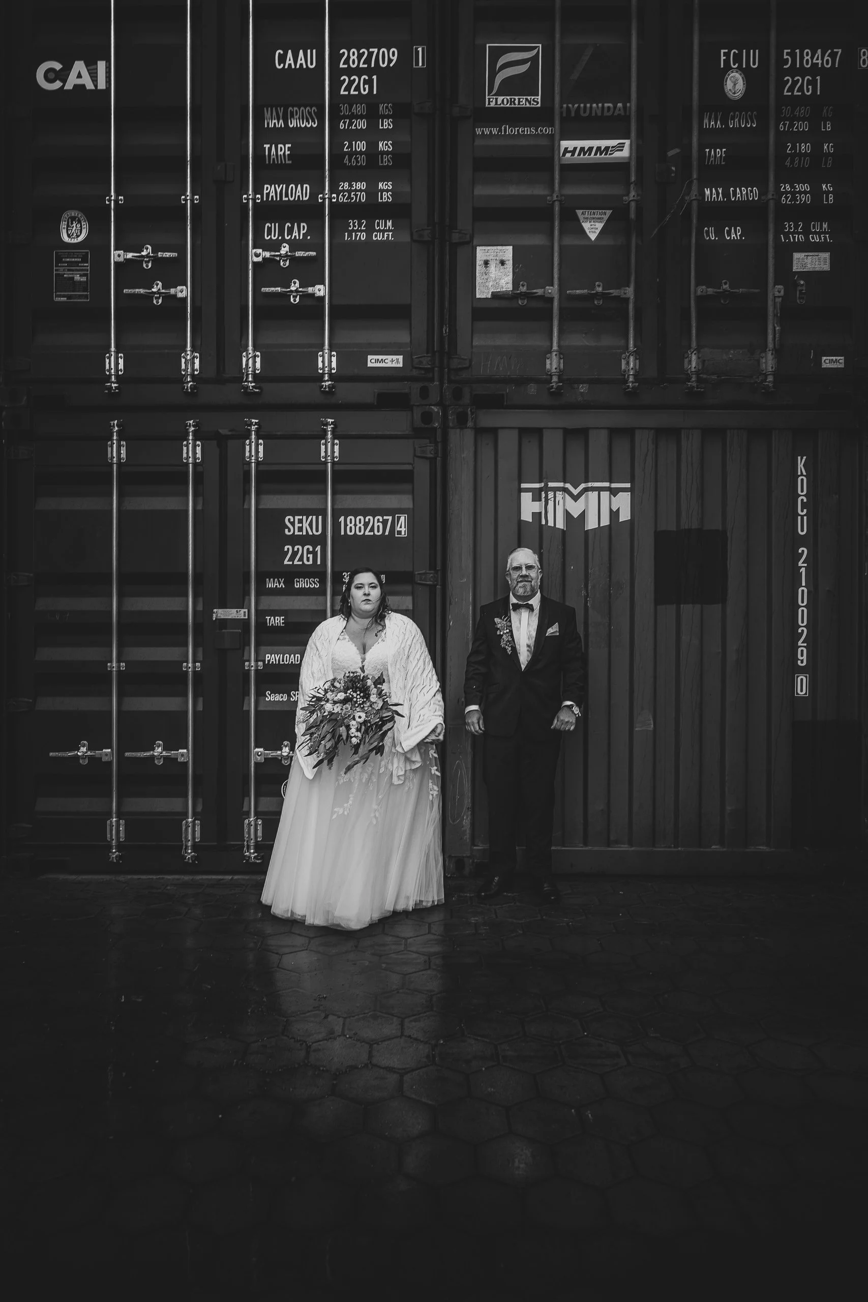 Ein Braut und ein Bräutigam stehen vor einem Container. Die Braut trägt ein langes Hochzeitskleid und hält einen Blumenstrauß, der Bräutigam trägt einen Anzug. Das Bild ist schwarz-weiß.