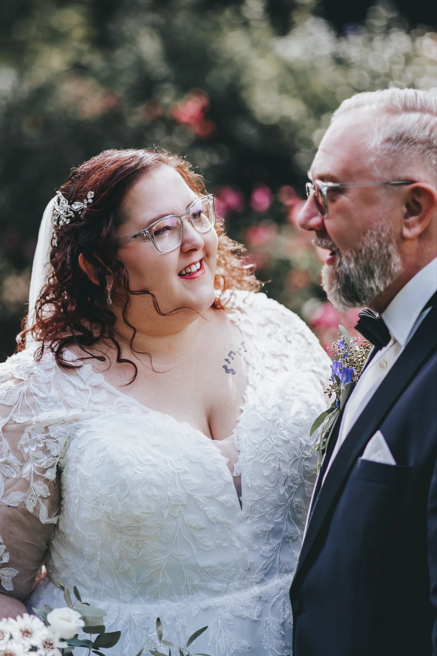 Ein Brautpaar bei einer Hochzeitszeremonie, die Braut trägt ein weißes Spitzenkleid und Brille, die beiden lächeln sich an.