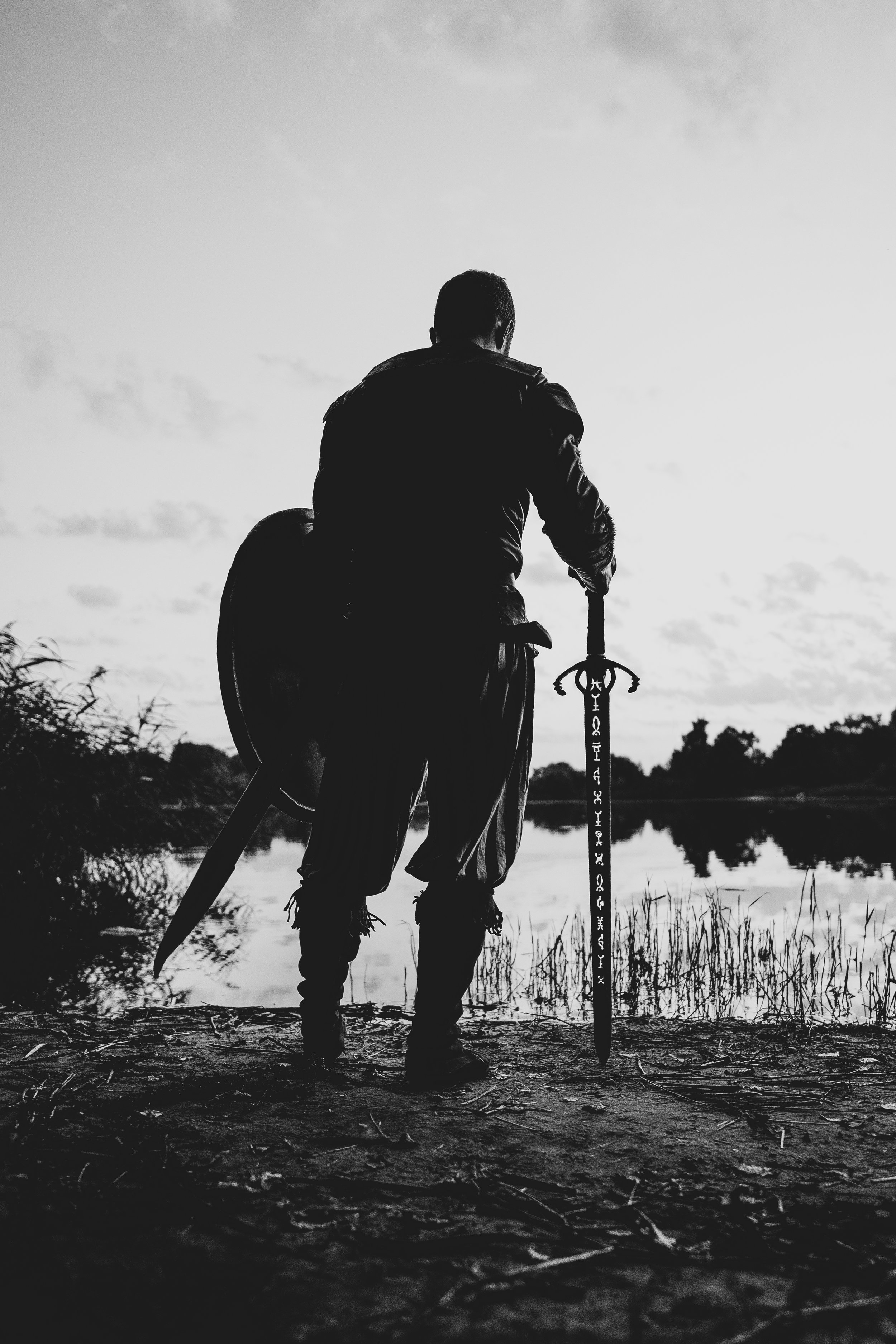 Silhouette eines Mannes im Piratenkostüm mit Schwert und Schild am Flussufer bei Sonnenaufgang oder Sonnenuntergang, schwarz-weiß