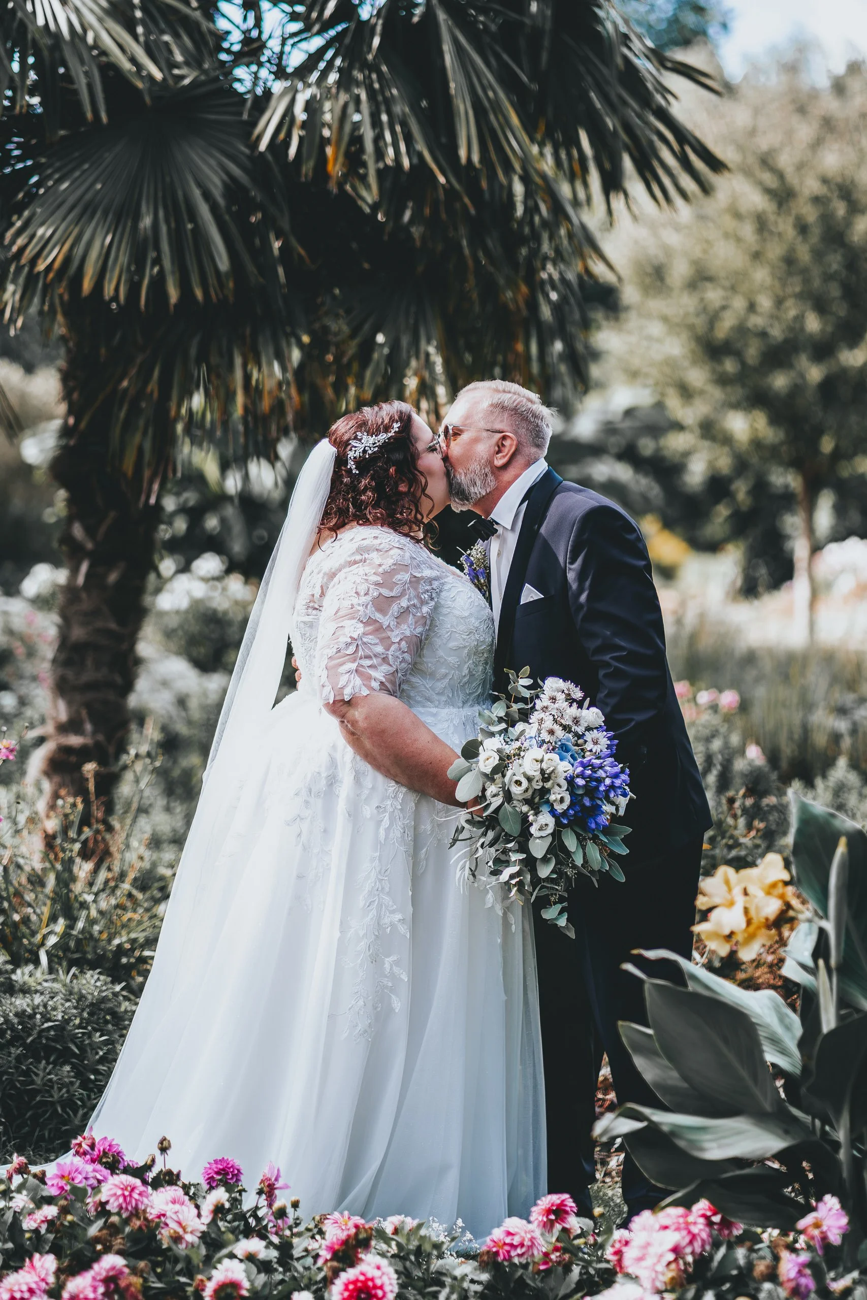 Ein Brautpaar küsst sich in einem Garten, die Braut trägt ein weißes Hochzeitskleid mit Spitze, hält einen Blumenstrauß in Blau, Weiß und Grün, und sie stehen vor einer Palme, umgeben von bunten Blumen.