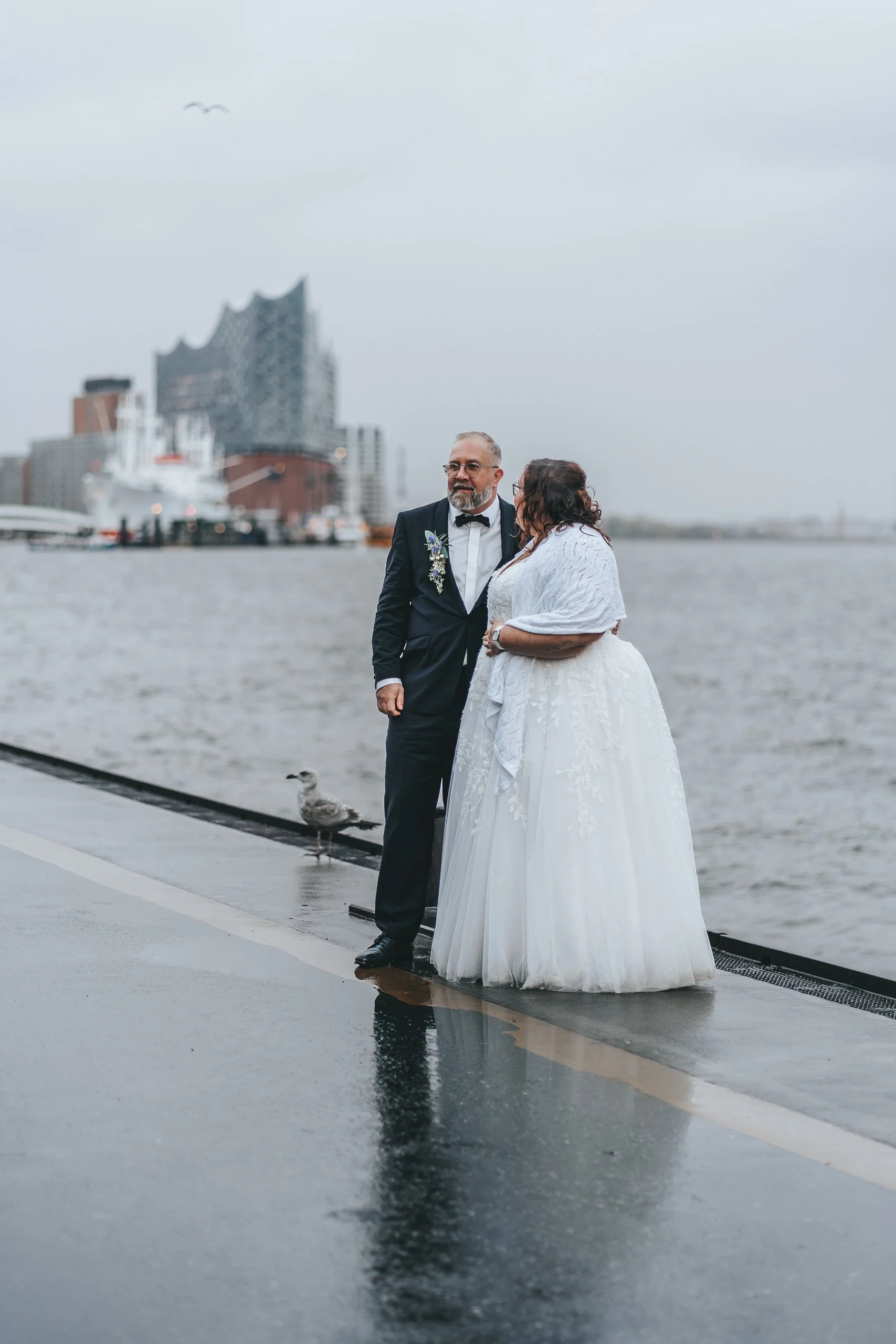 Ein brautpaar mit Gummistiefeln steht am Wasser auf einem Pier, im Hintergrund die Elbphilharmonie in Hamburg, ein Grauer Himmel und Wasser