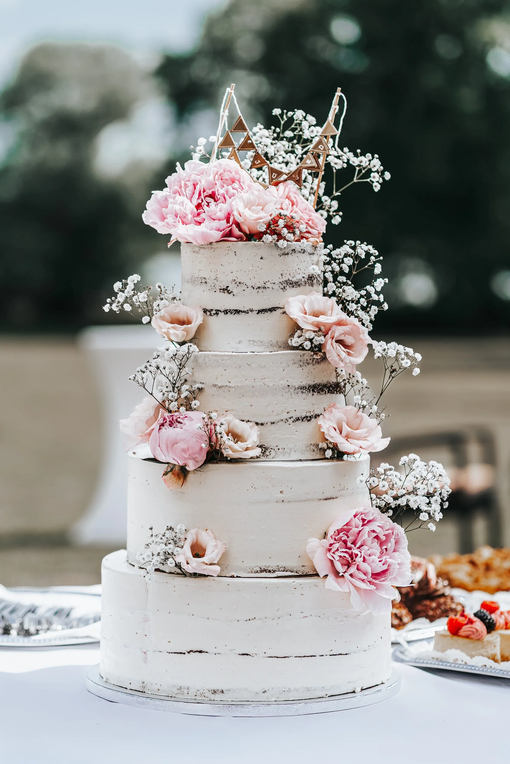 Mehrstöckige weiße Hochzeitstorte im naked-cake-style mit rosa und weißen Blumen und einer kleinen Dekoration auf Top, auf einem weißen Tisch, im Hintergrund unscharfe Natur.