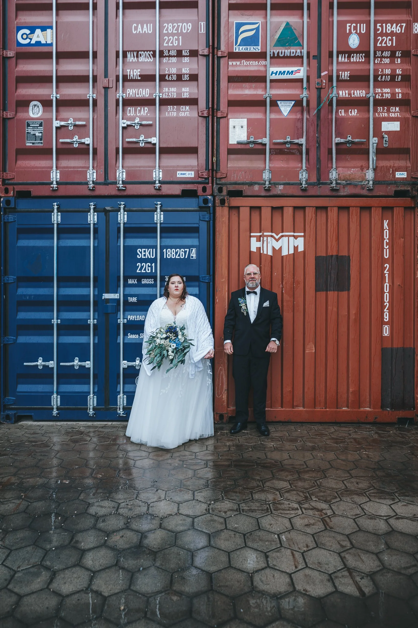 Ein Brautpaar in formeller Kleidung steht vor bunte Container, der Boden ist nass, im Hintergrund sind mehrere Container in Rot, Blau und Grün.