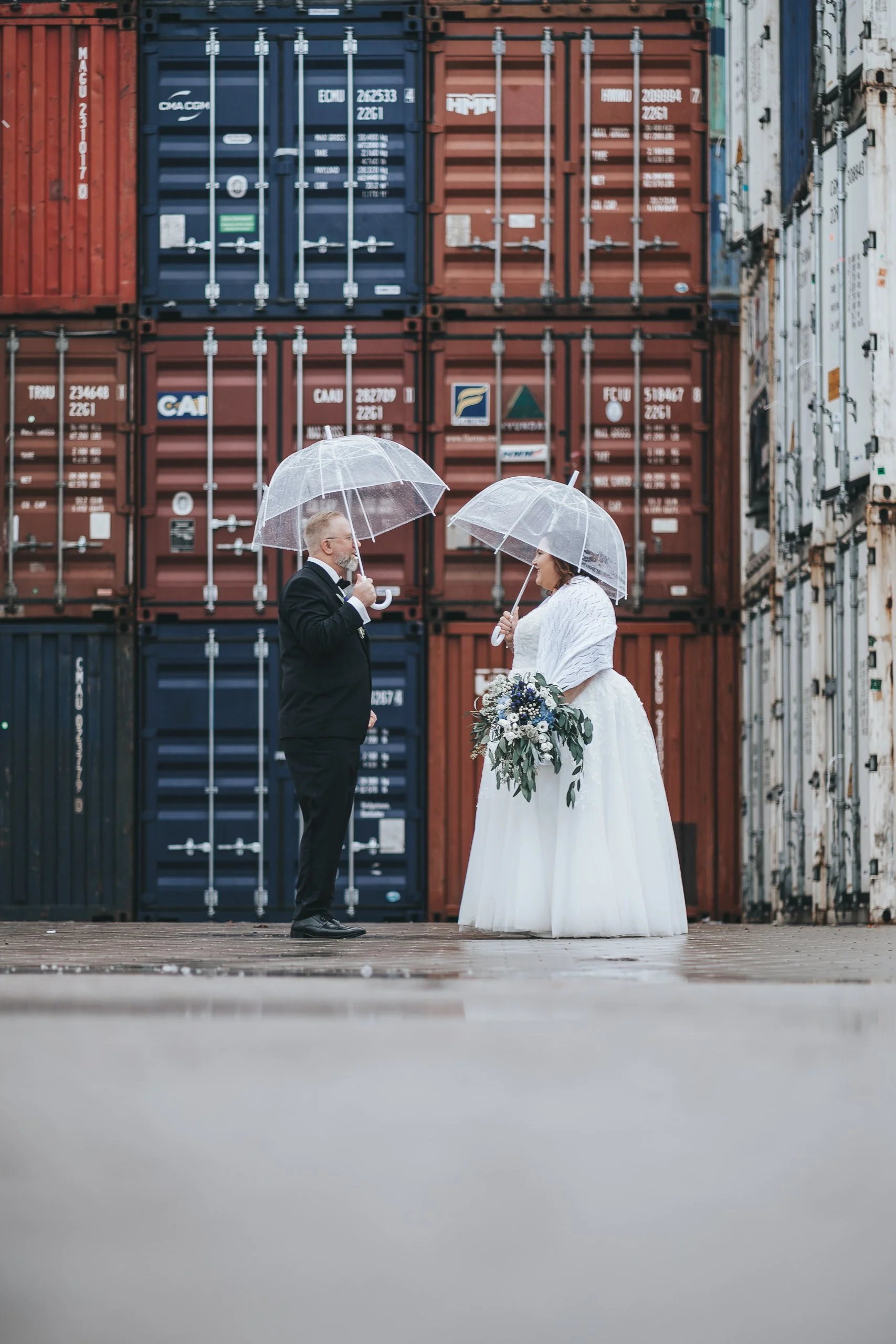 Ein heiratendes Paar in Hochzeitskleidung unter transparenten Regenschirmen vor einem Stapel von Industriecontainern.