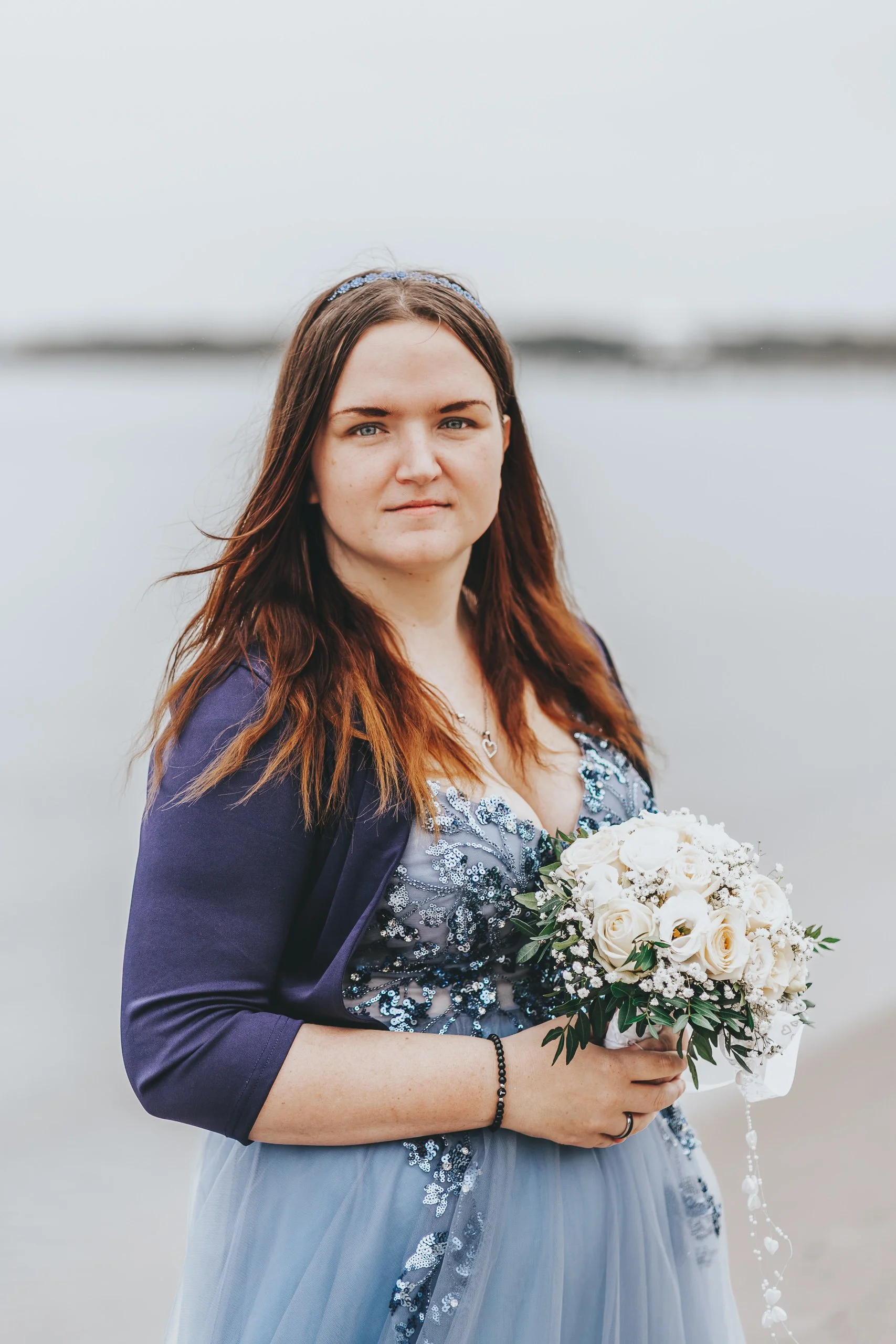 Eine junge Frau hält einen Brautstrauß mit weißen Rosen und grünen Blättern, trägt ein dunkelblaues Kleid mit aufwendigen Stickereien, steht am Wasser bei bewölktem Himmel.