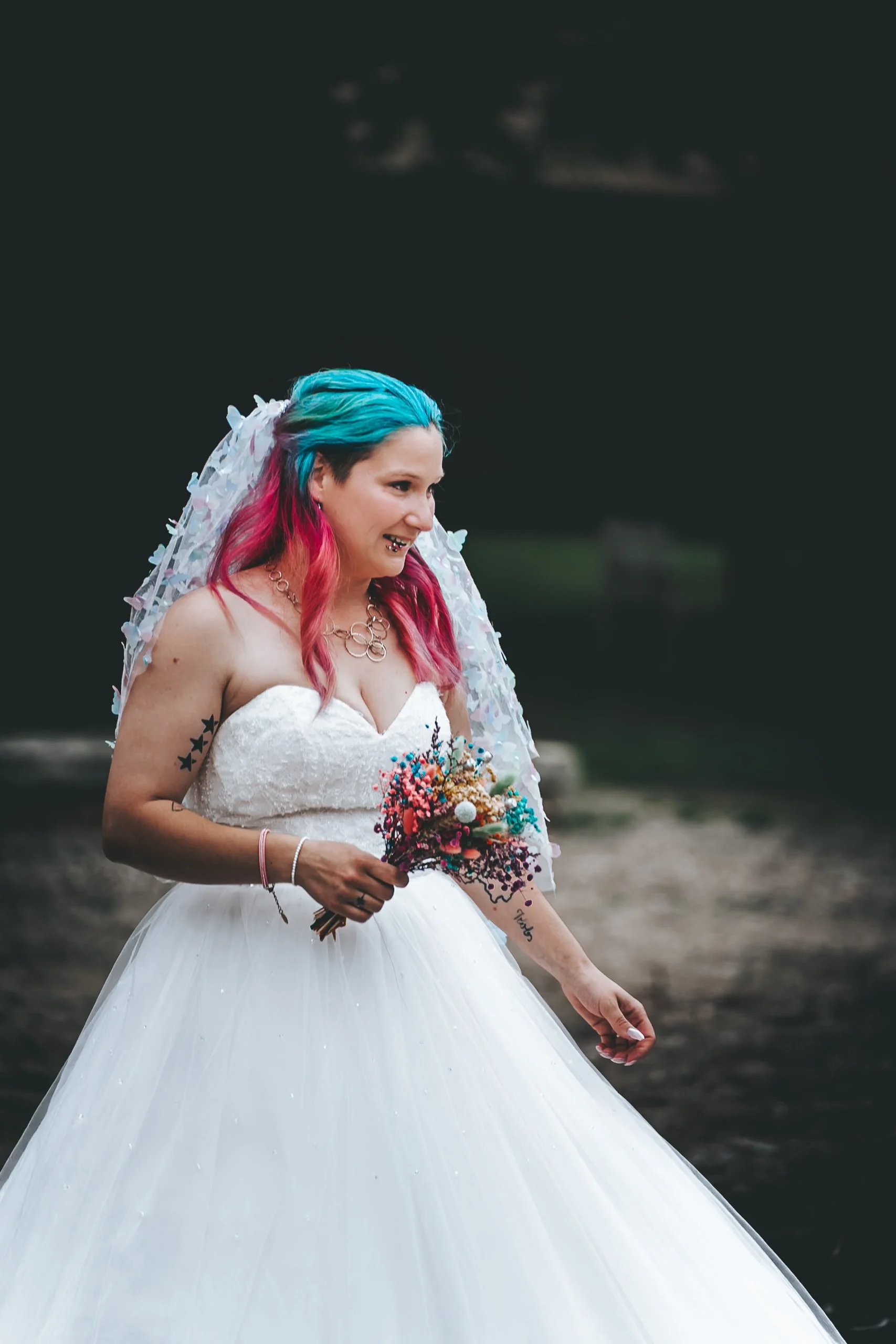 Eine Frau in einem Brautkleid mit buntem Haar hält einen Blumenstrauß und lächelt.