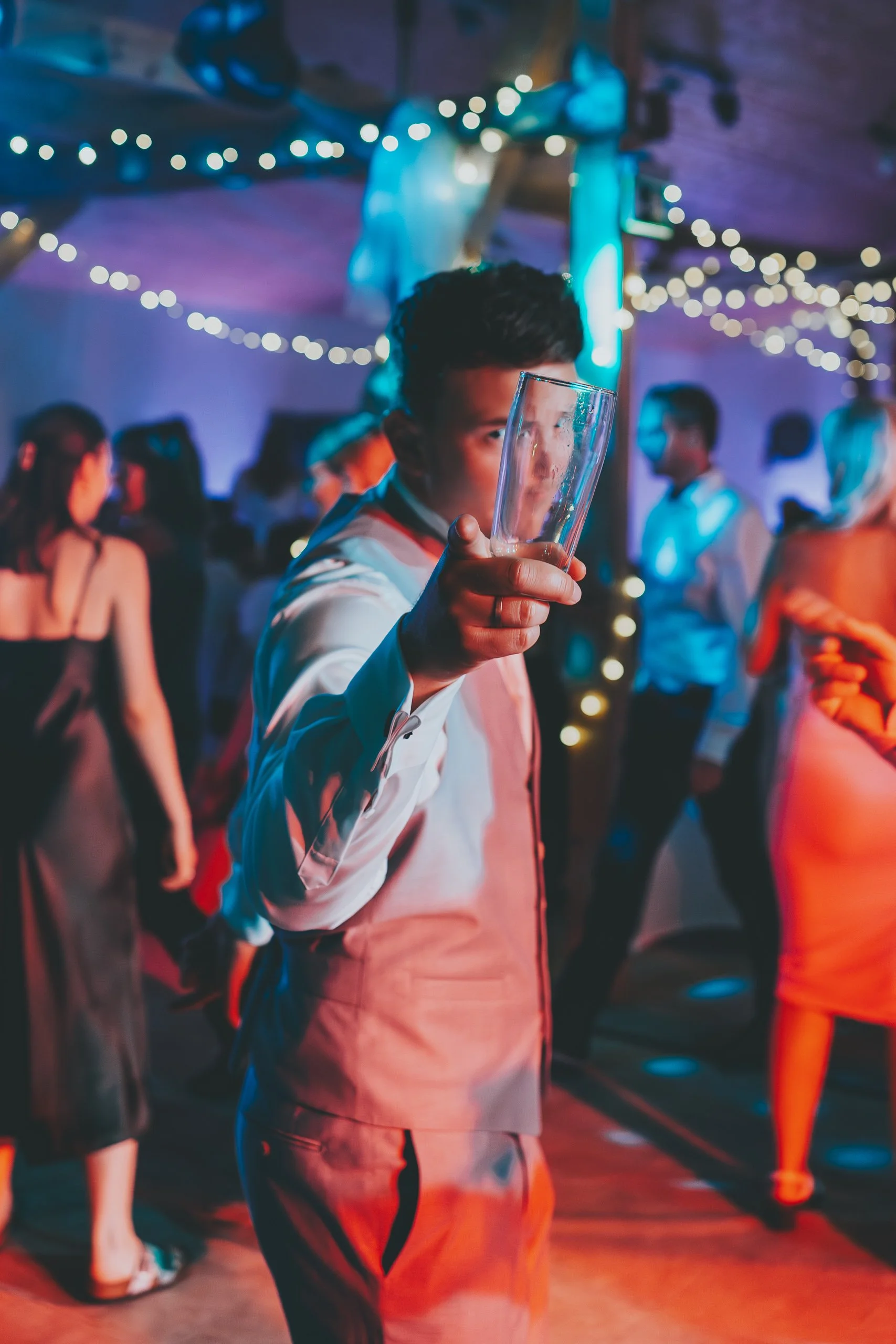 Junge in Hemd und Weste auf einer Party in einem Raum mit Lichterdekoration und mehreren Menschen, hält ein leeres Glas in die Kamera.