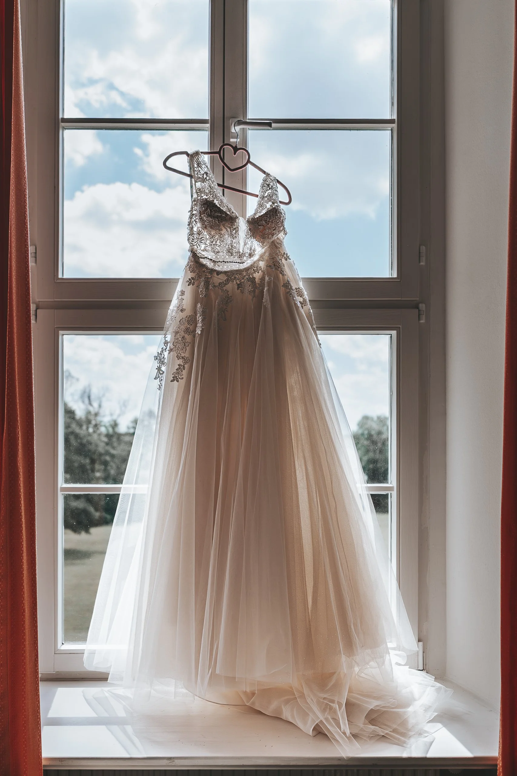 Elegantes Brautkleid mit Spitzen-Details hängt an einem Herz-Haken vor einem großen Fenster mit Blick auf den Himmel.
