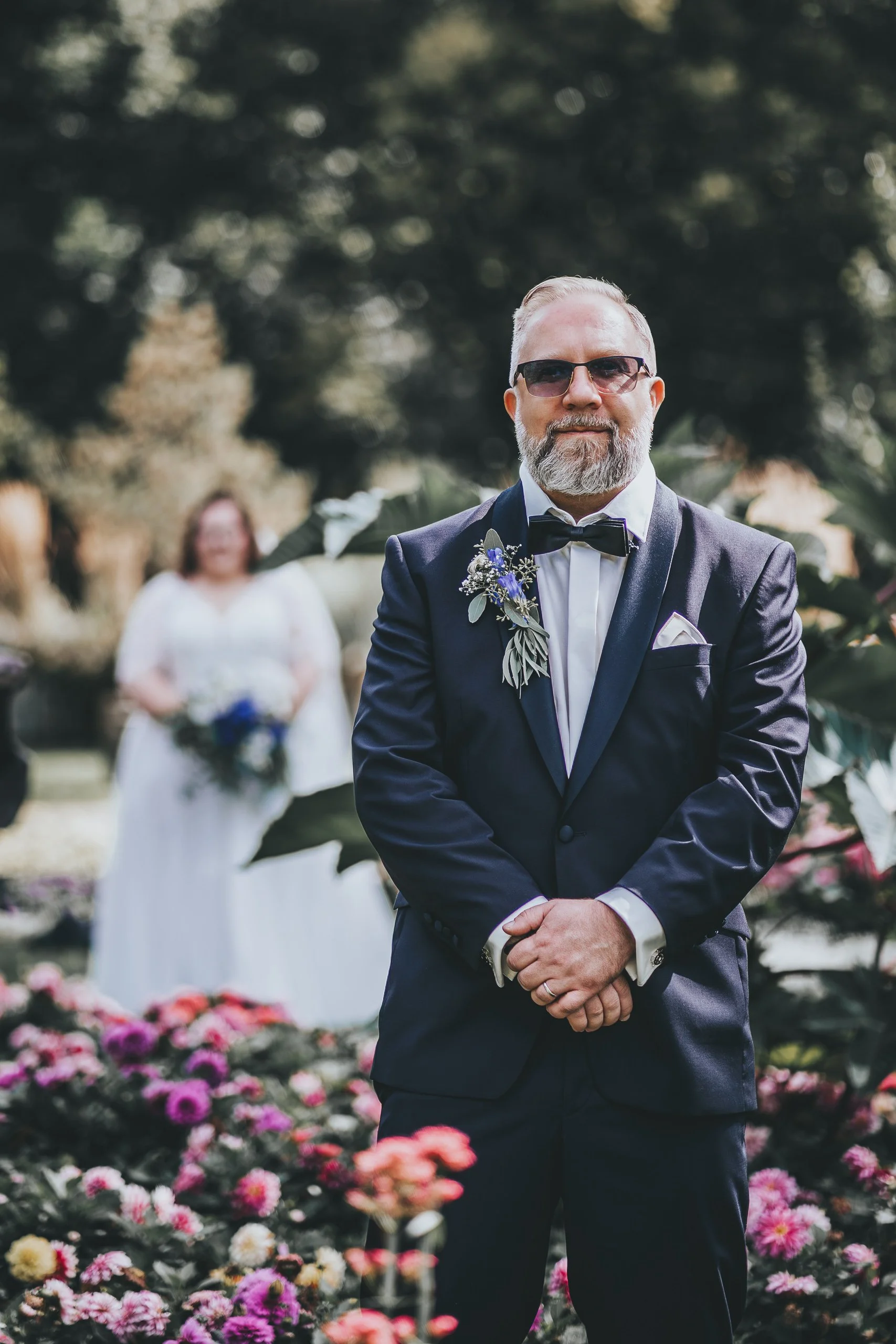 Ein Mann in formeller Kleidung, schwarzen Anzug mit Fliege und Sonnenbrille, steht vor einer Gruppe von Menschen bei einer Hochzeit, im Hintergrund ein Blumenbogen und eine blühende Gartenlandschaft.