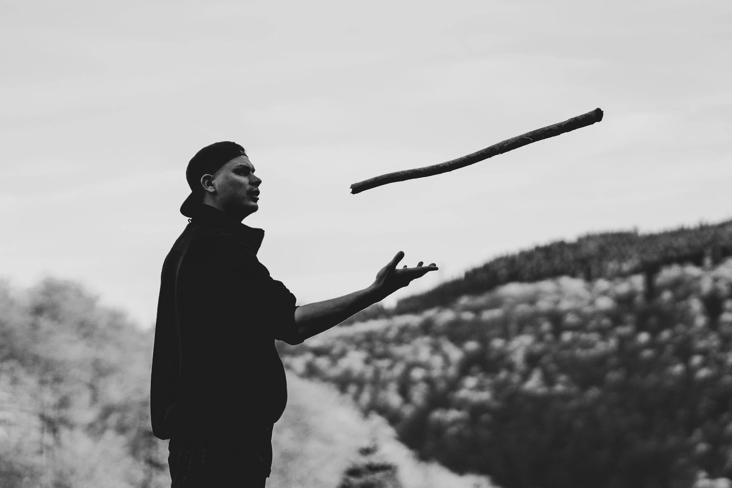Ein junger Mann wirft einen Stock in einer ländlichen Umgebung, im Hintergrund sind Hügel und Bäume. Das Bild ist Schwarzweiß.