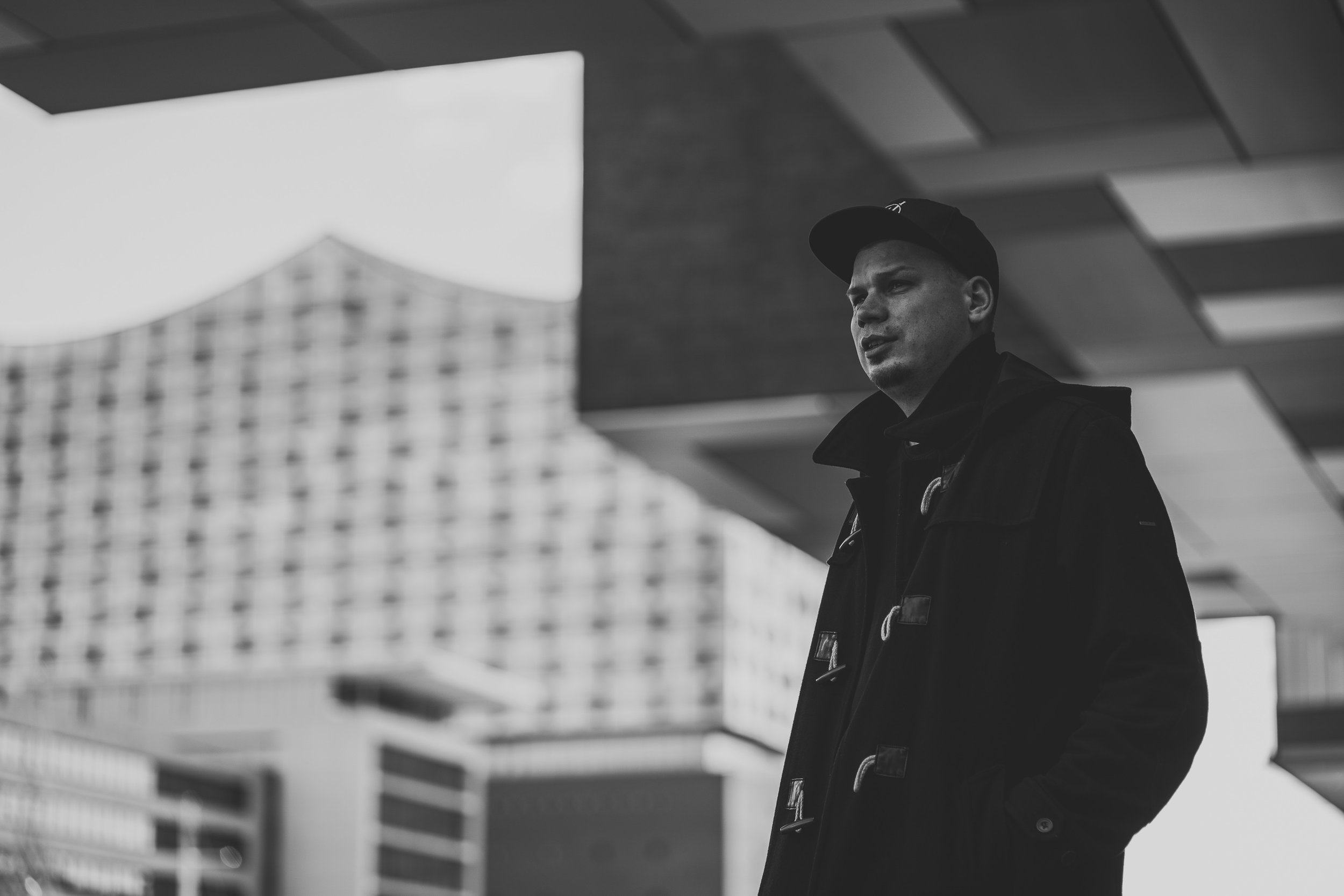 Ein junger Mann trägt eine dunkle Jacke und eine schwarze Baseballkappe, steht im Freien vor einem modernen Gebäudekomplex, schwarz-weiß Aufnahme.