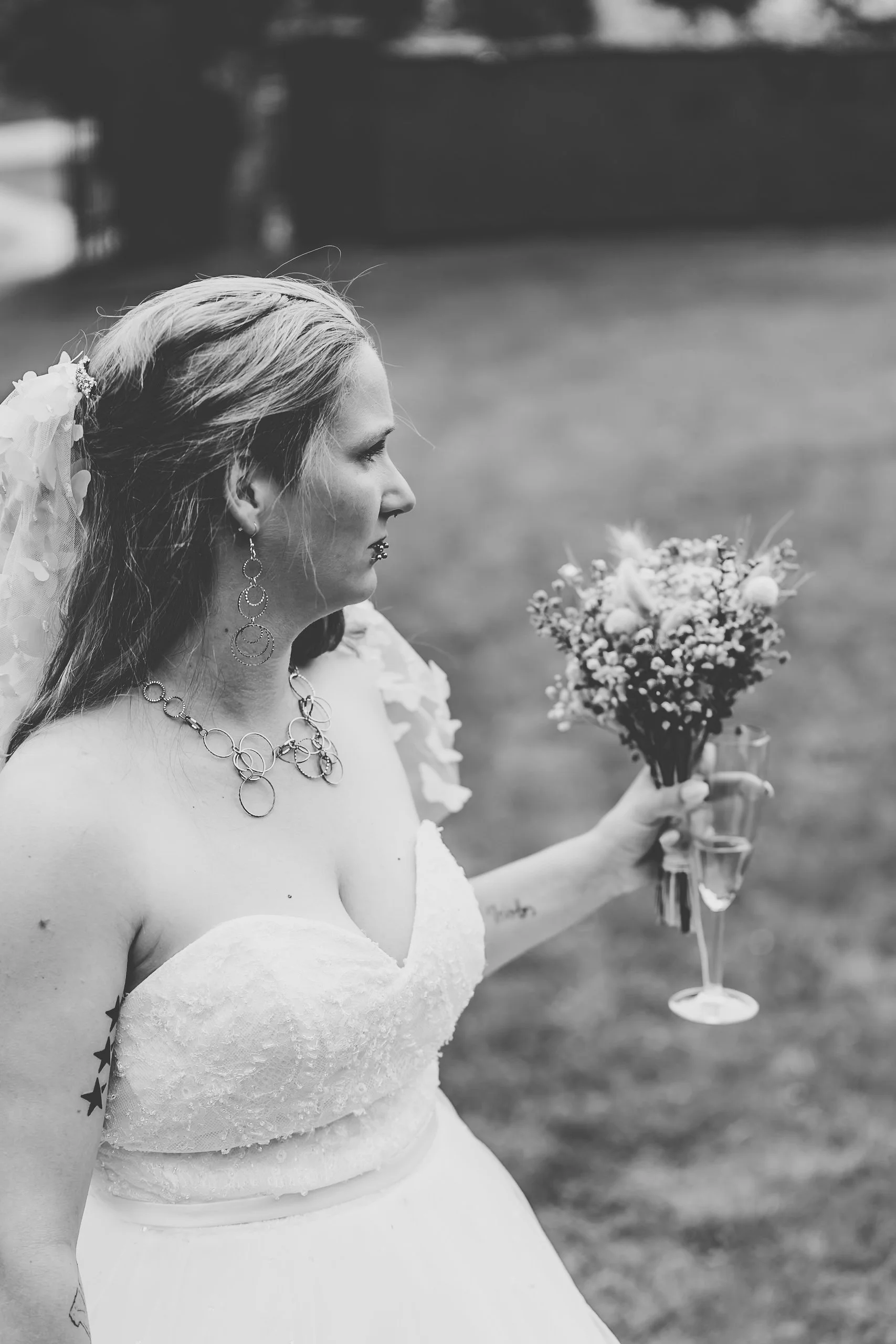 Eine Braut in Brautkleid mit Schmuck hält ein Blumenbouquet und ein Glas Sekt im Freien.