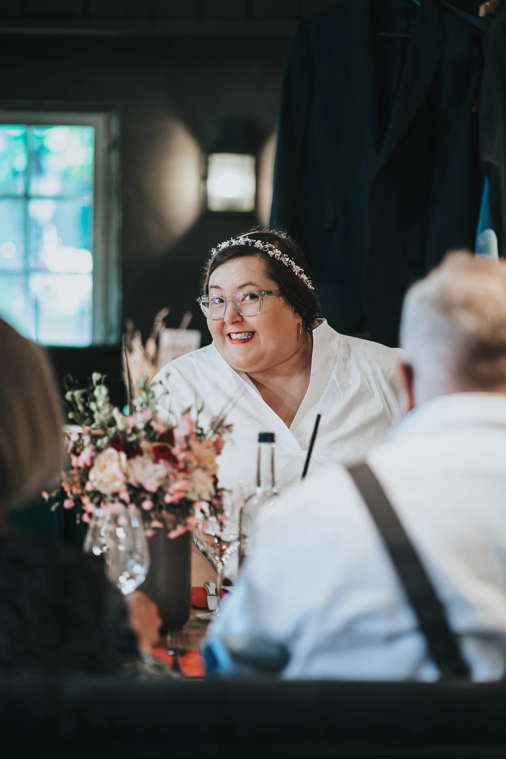Eine lachende Frau mit Brille und Diadem bei einem Treffen oder einer Feier, umgeben von anderen Personen und einer Vase mit Blumen auf dem Tisch.