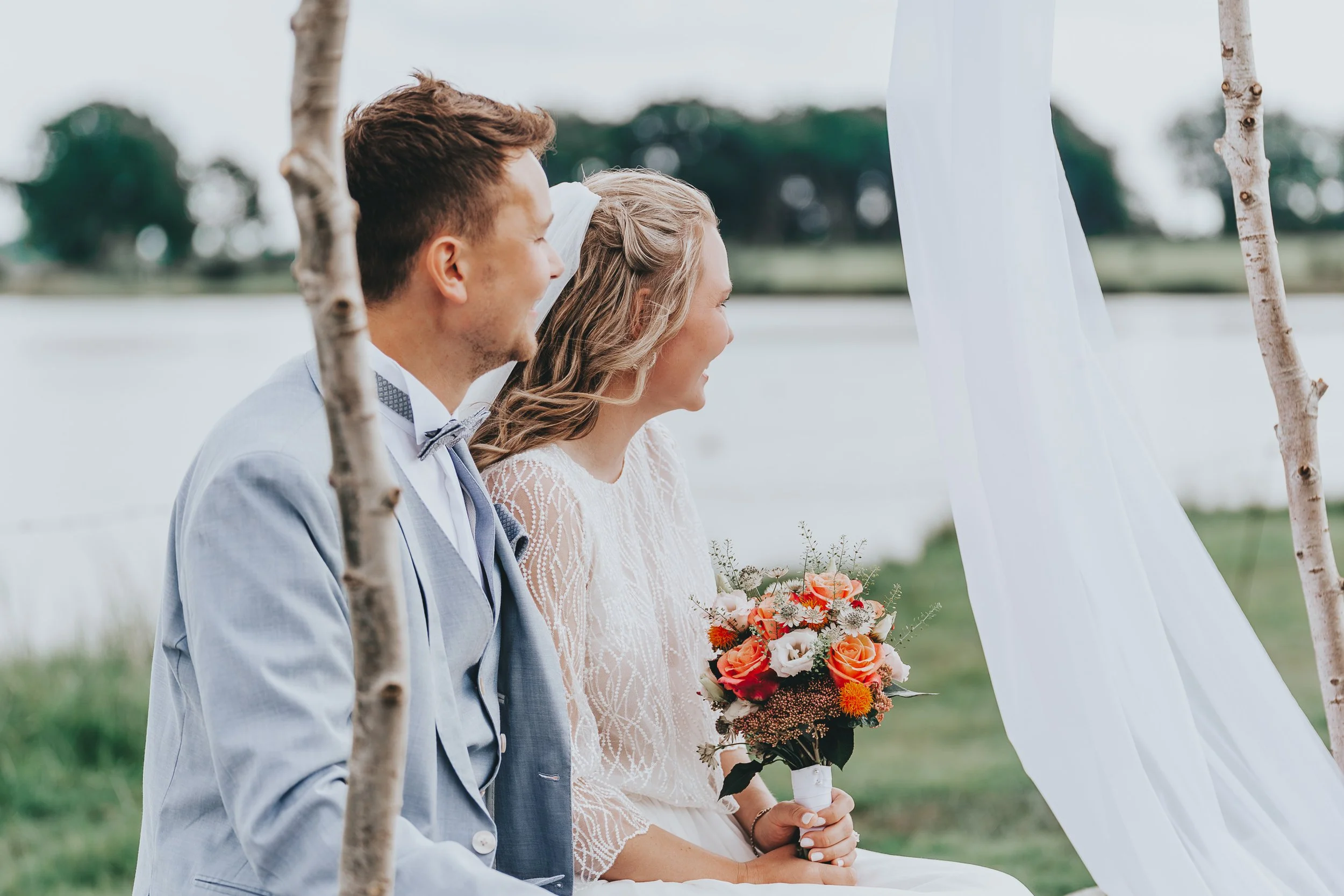 Braut und Bräutigam sitzen bei ihrer Hochzeit im Freien, umgeben von weißen Stoffen und Birkenstämmen. Die Braut hält einen bunten Blumenstrauß. Sie lachen und freuen sich, während sie an einem Gewässer im Hintergrund vorbeischauen.