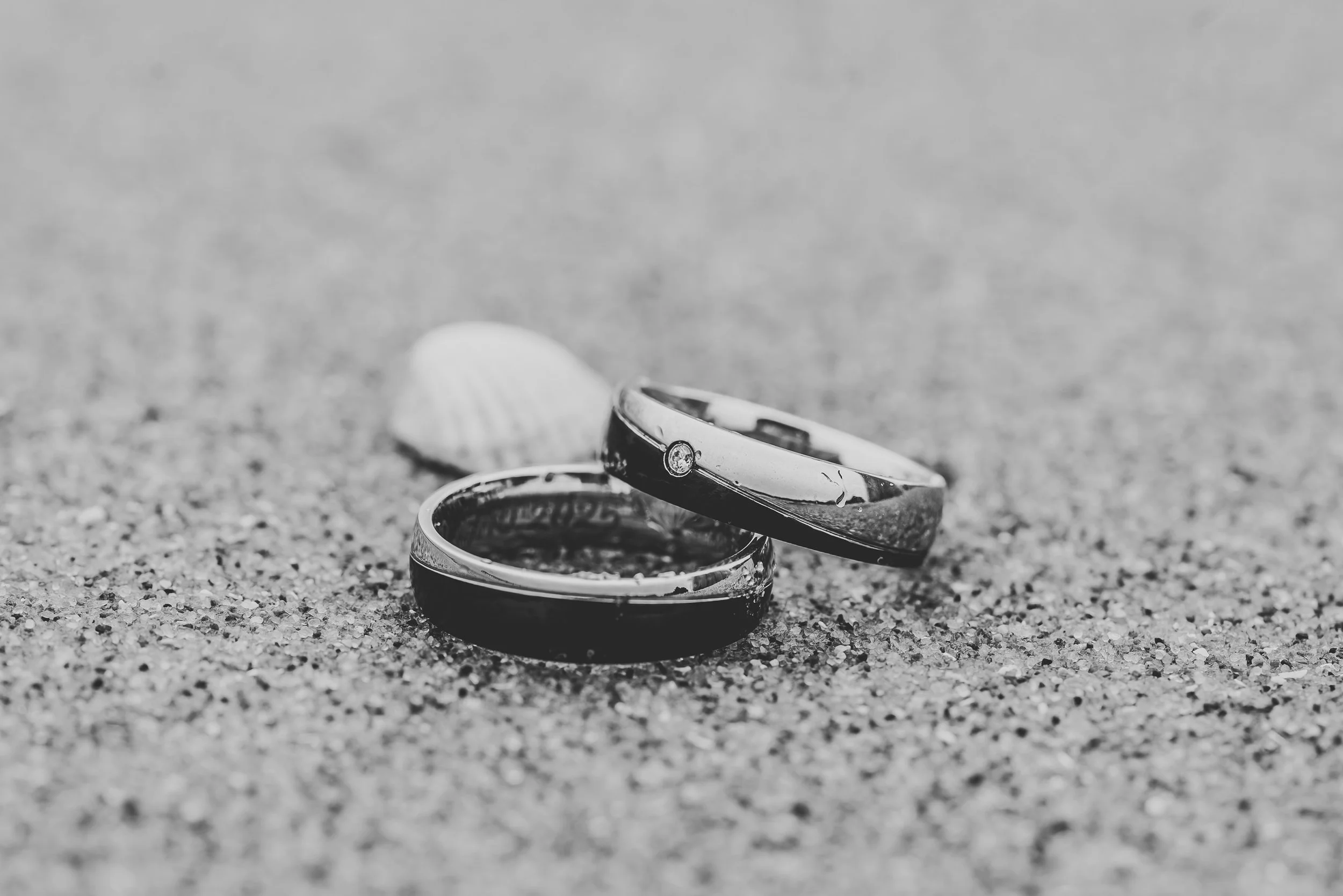 Zwei Eheringe auf Sand mit Muscheln im Hintergrund, schwarz-weiße Aufnahme.