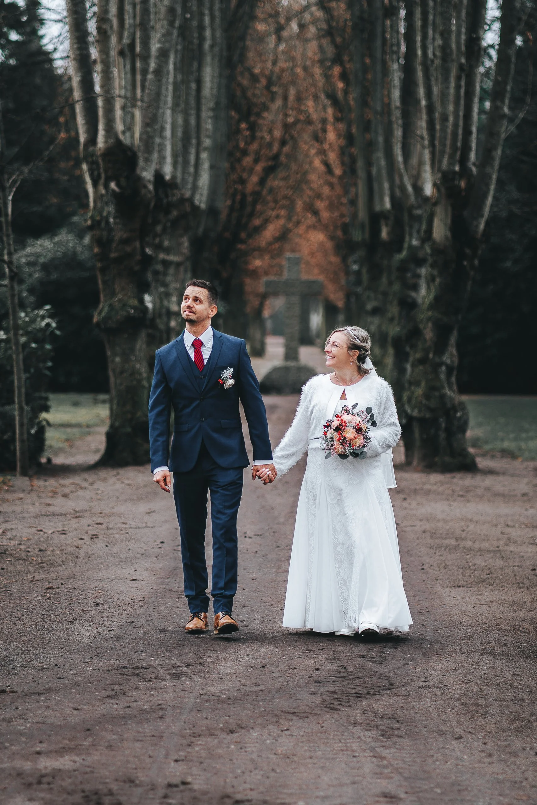 Ein Paar in Hochzeitskleidung läuft Hand in Hand durch einen Park mit großen Bäumen und einem Kreuz im Hintergrund.