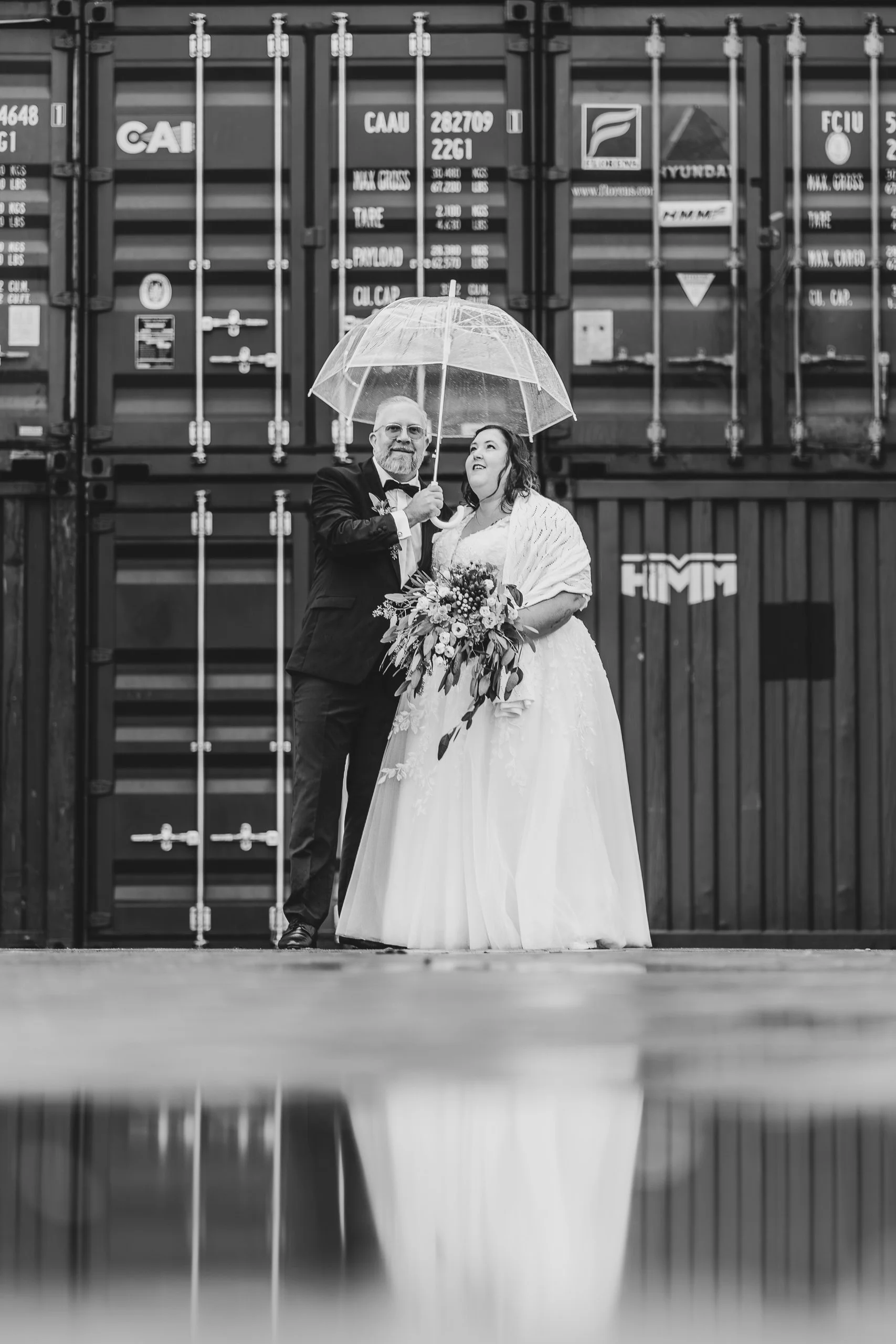 Braut und Bräutigam im Regen unter transparentem Regenschirm vor einem Container