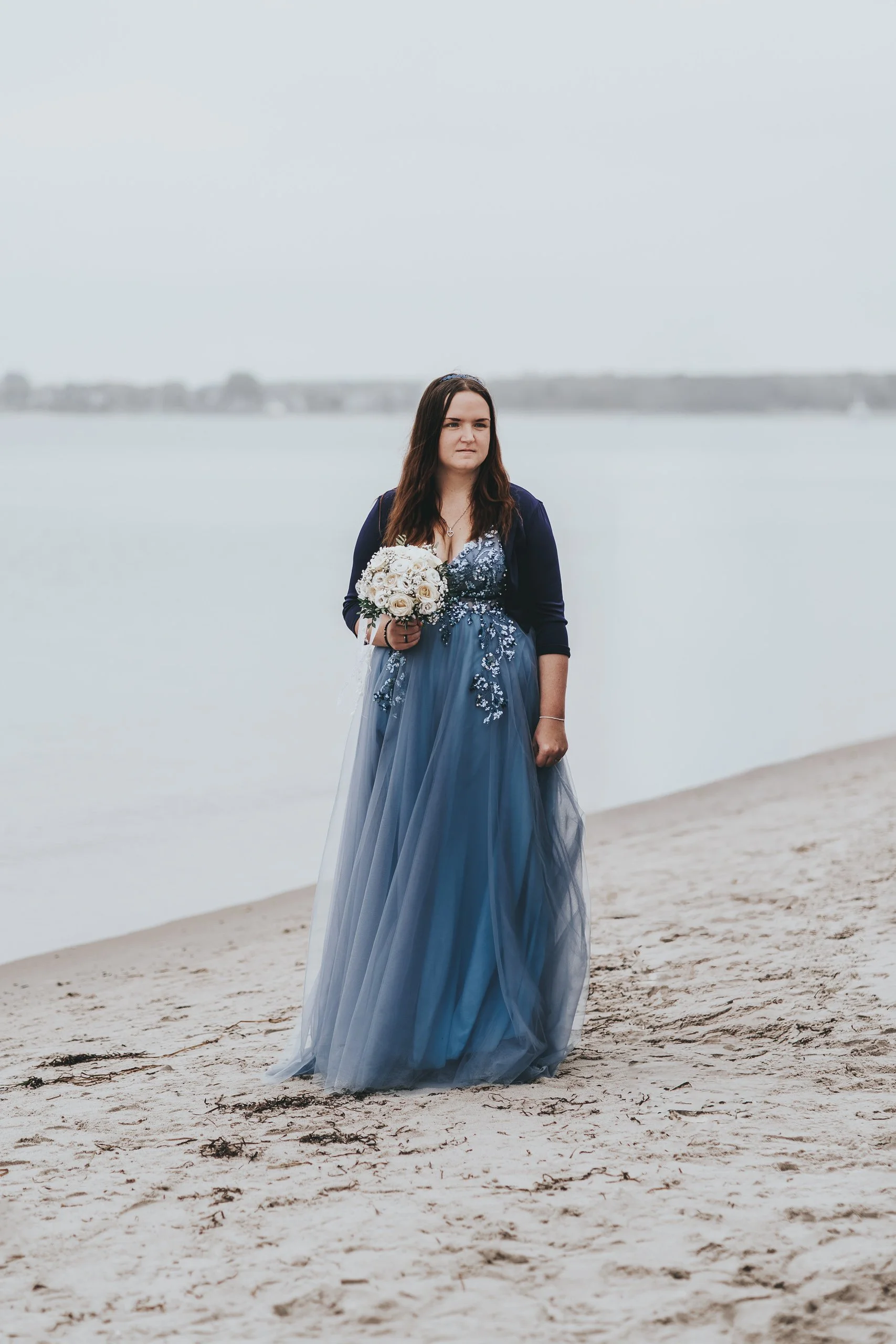 Eine Frau in einem blauen Abendkleid mit Blumenstickereien hält einen Blumenstrauß und steht am Strand.