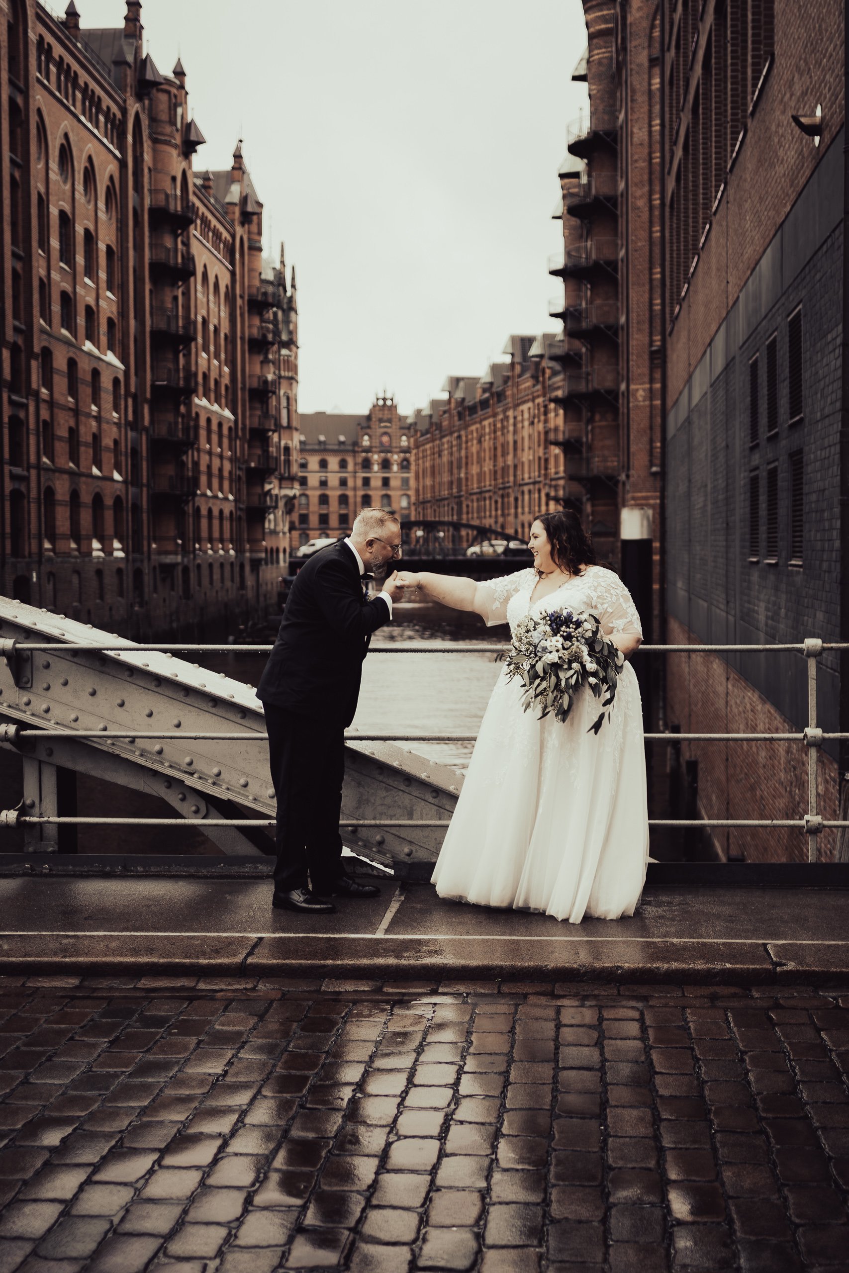 Ein Mann und eine Frau in Hochzeitskleidung stehen auf einer Brücke in einer Stadt in einer romantischen Geste. Der Mann küsst die Hand der Frau, die ein Brautkleid trägt und eine Blumenstrauß hält. Im Hintergrund sind historische Backsteingebäude en