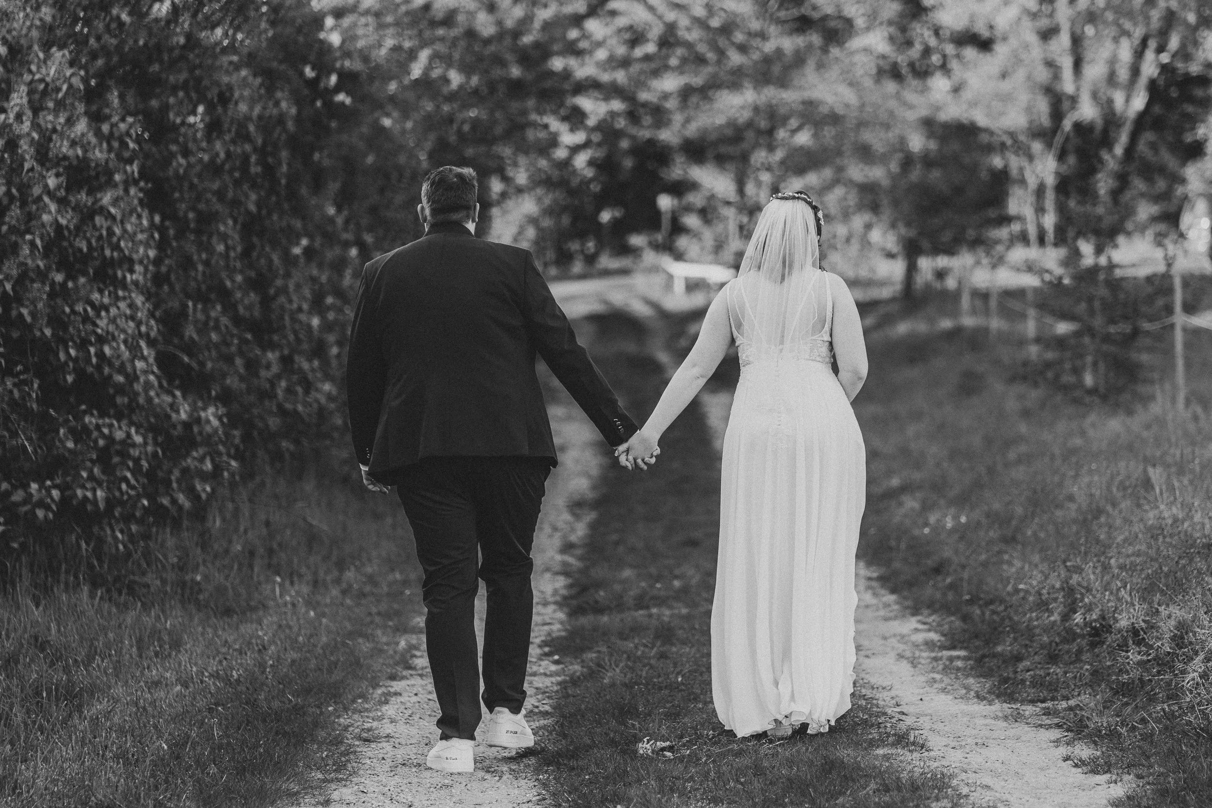 Ein Brautpaar läuft Hand in Hand auf einem schmalen Pfad durch eine grüne Parklandschaft. Es ist schwarz-weiß fotografiert, der Hintergrund ist mit Bäumen und Blättern gefüllt.