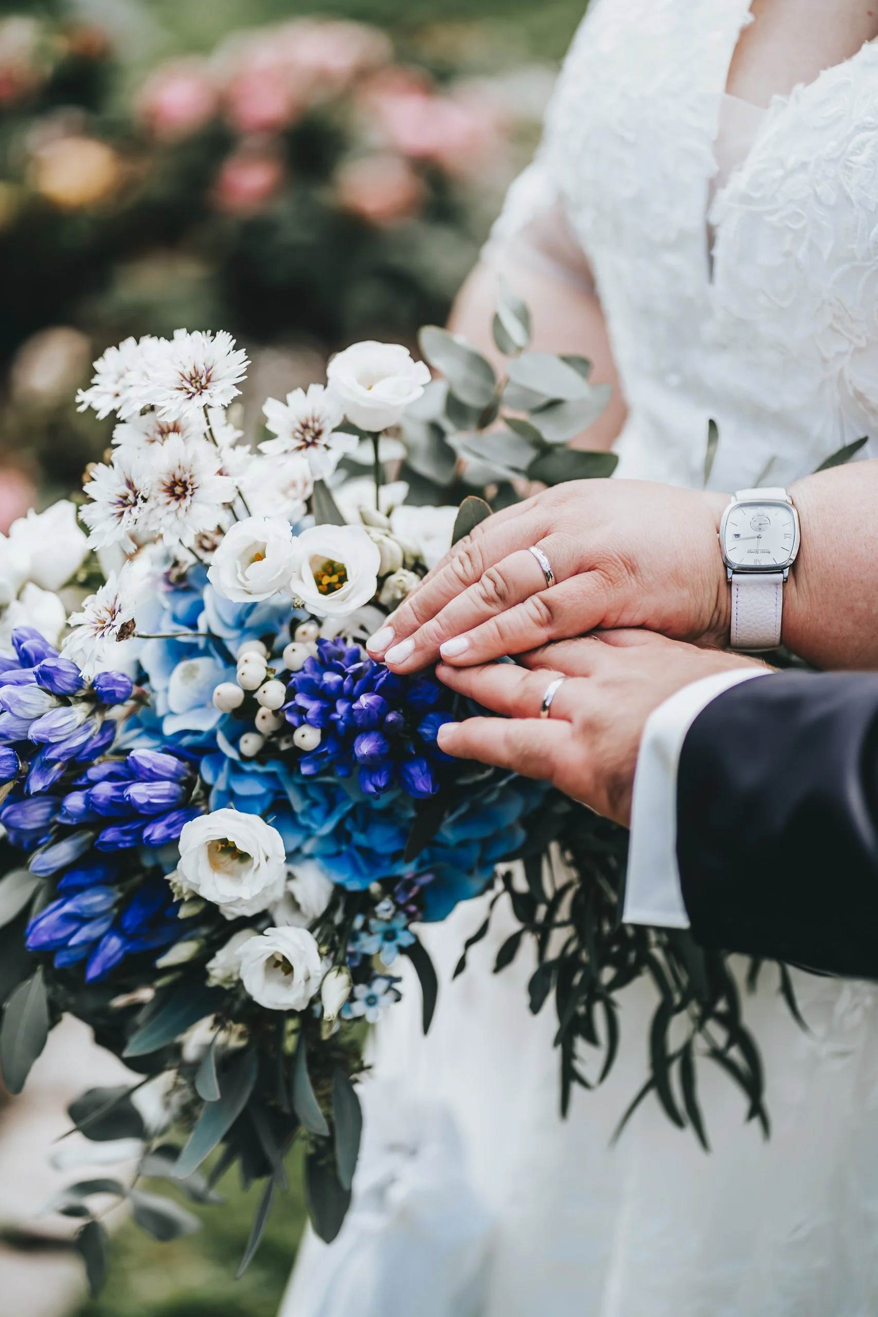 Hochzeitspaar hält Blumenstrauß mit weißen, blauen und violetten Blumen, nahe bei den Händen, die Eheringe tragen, im Fokus.