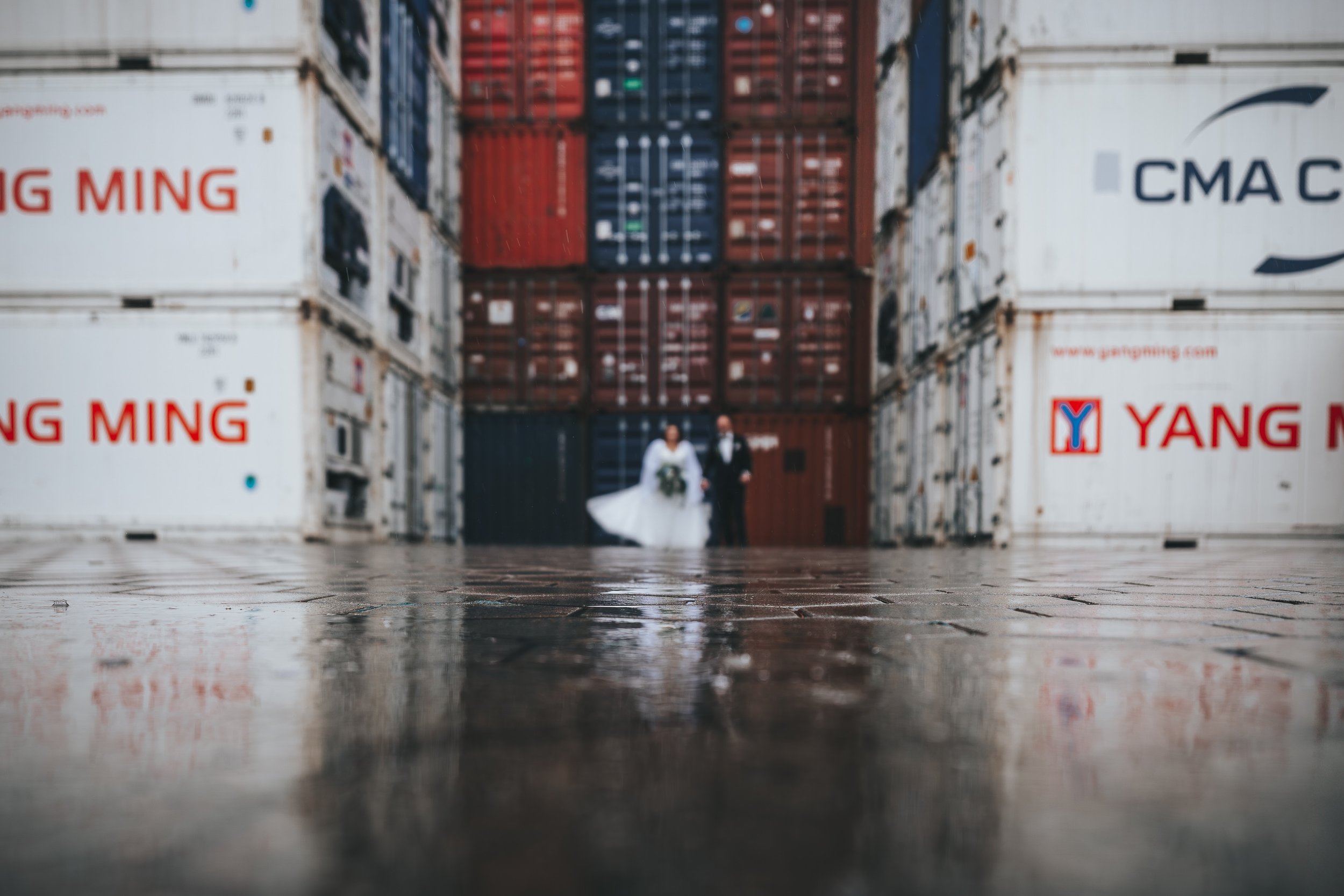 Verzerrtes Foto eines Brautpaares in Hochzeitskleidung, das auf einer Fabrikhalle mit großen Container-Lagerhäusern im Hintergrund steht.