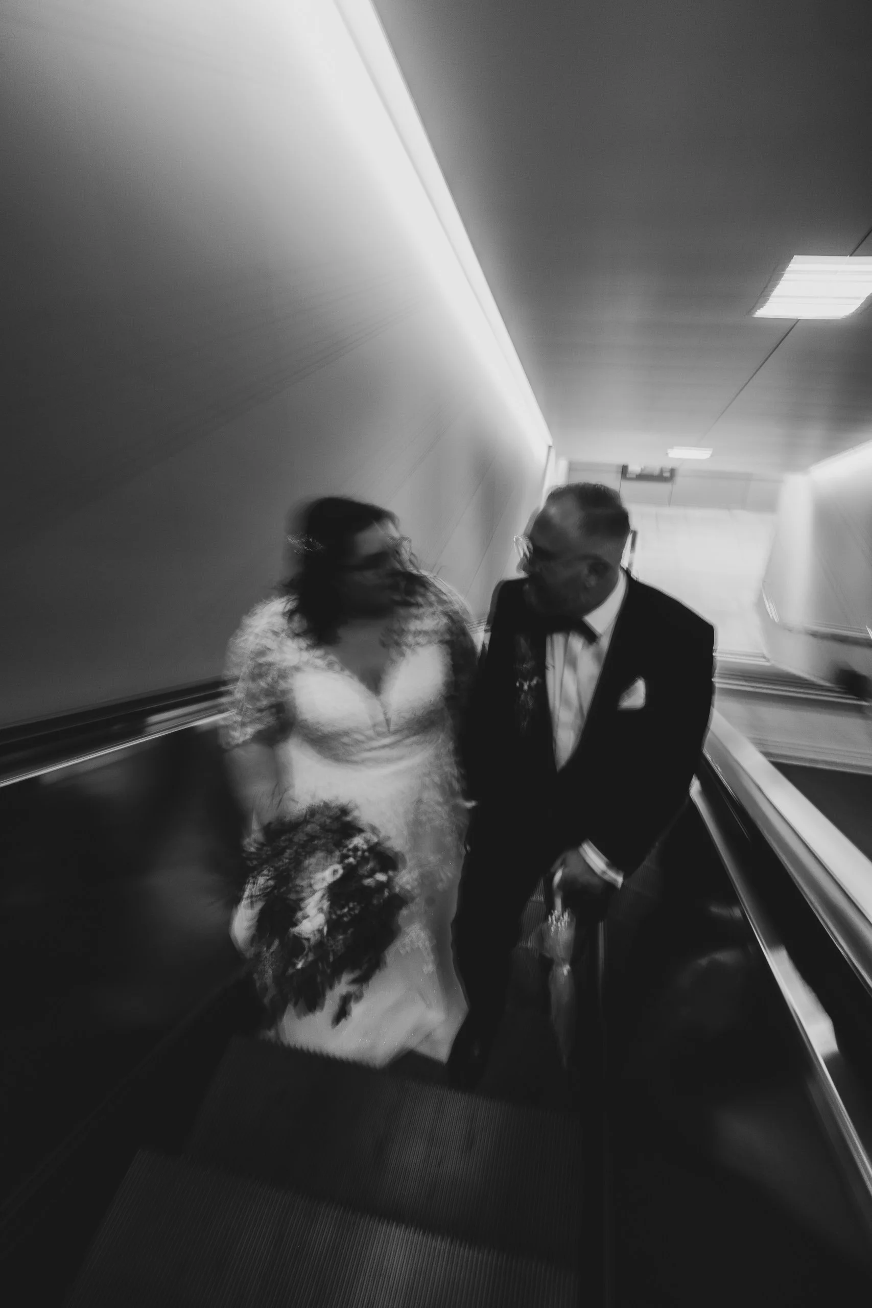 Ein hochzeitsfoto von einem Brautpaar auf einer Rolltreppe, schwarz-weiß, gestellte Aufnahme, unscharf, von oben fotografiert.