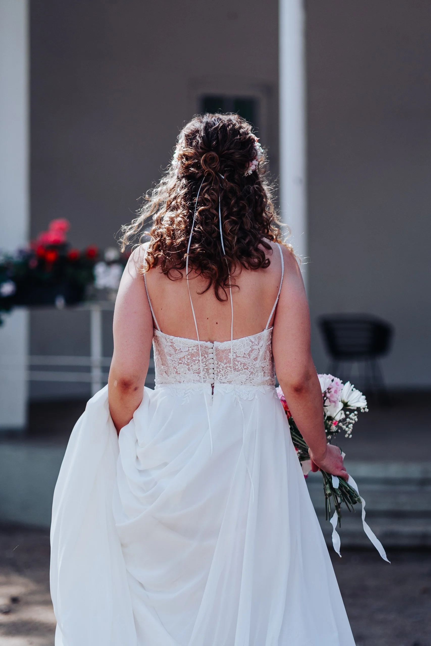Braut von hinten im weißen Kleid mit langen Locken und Blumenstrauß.
