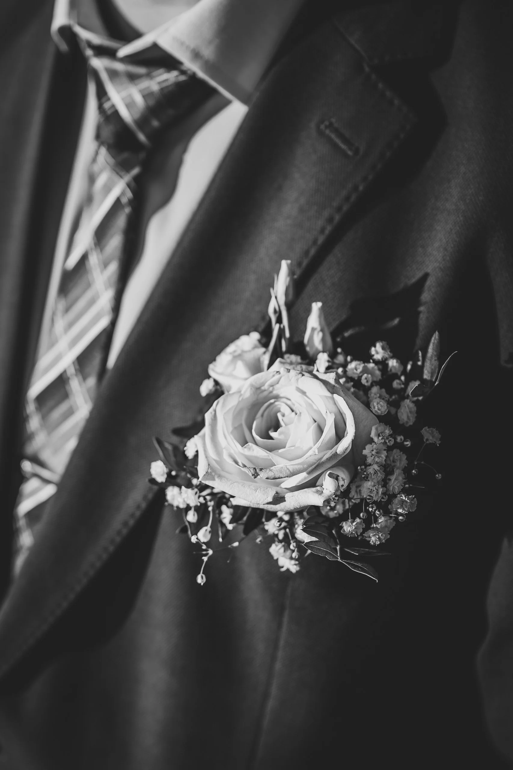 Schwarz-Weiß-Foto eines Anzugs mit Fliege und einer Ansteckblume, bestehend aus einer Rose und kleinen Blumen, an der Jacke. 
