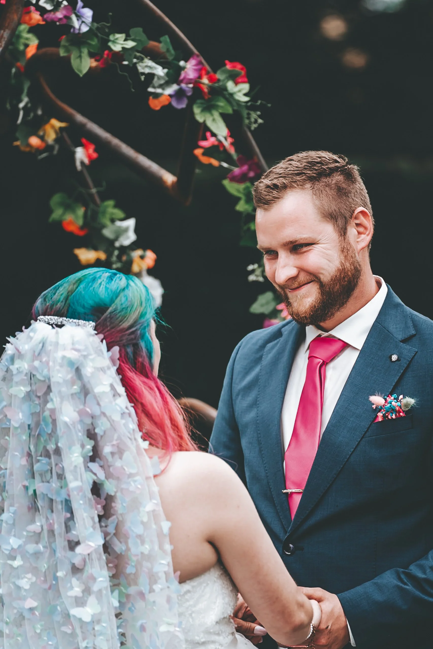 Ein Paar bei einer Hochzeitszeremonie, der Mann trägt einen blauen Sakko, eine weiße Hemd und eine rosa Krawatte, die Frau hat bunte Haare und trägt ein Brautkleid mit einer Schleierhaube. Im Hintergrund sind Blumen und ein dekorativer Bogen sichtbar
