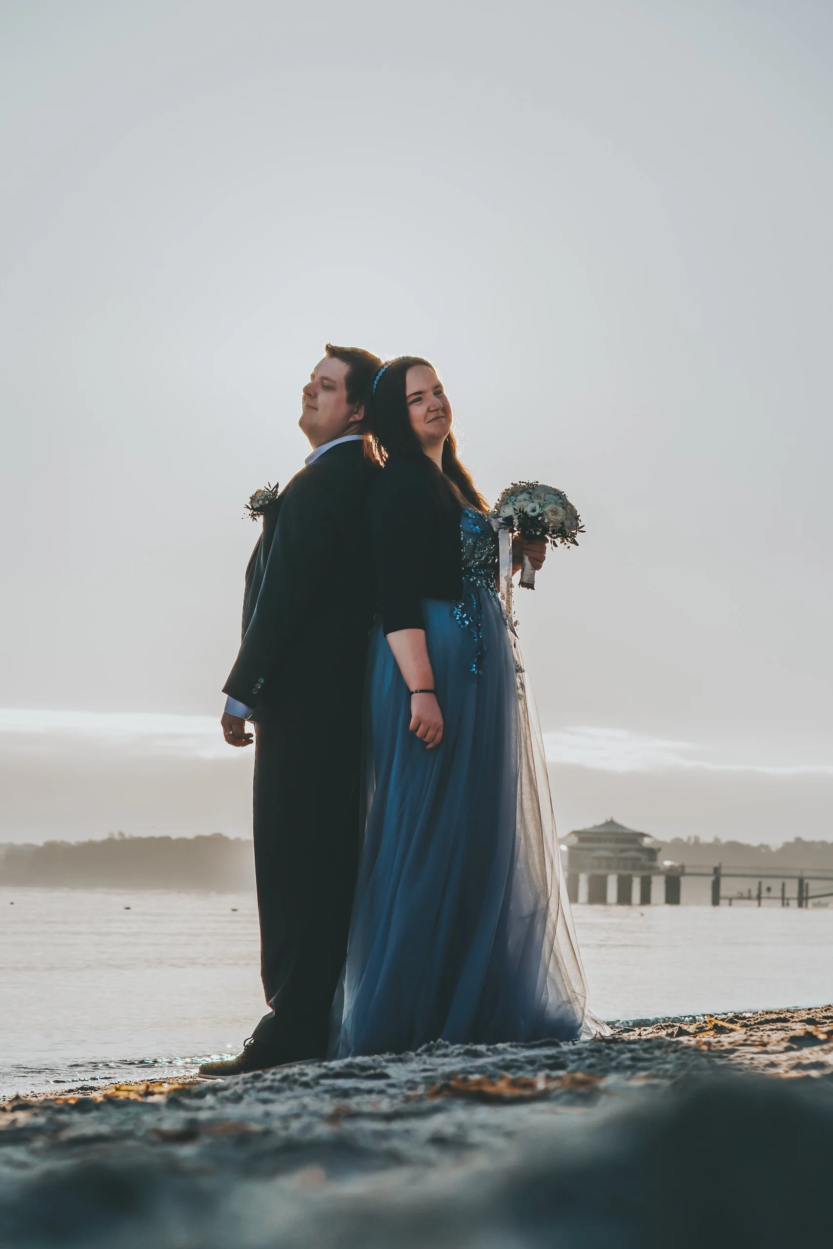 Braut und Bräutigam stehen an einem Strand im Sonnenuntergang, der Bräutigam trägt einen schwarzen Anzug, die Braut ein blaues Kleid und hält einen Blumenstrauß.