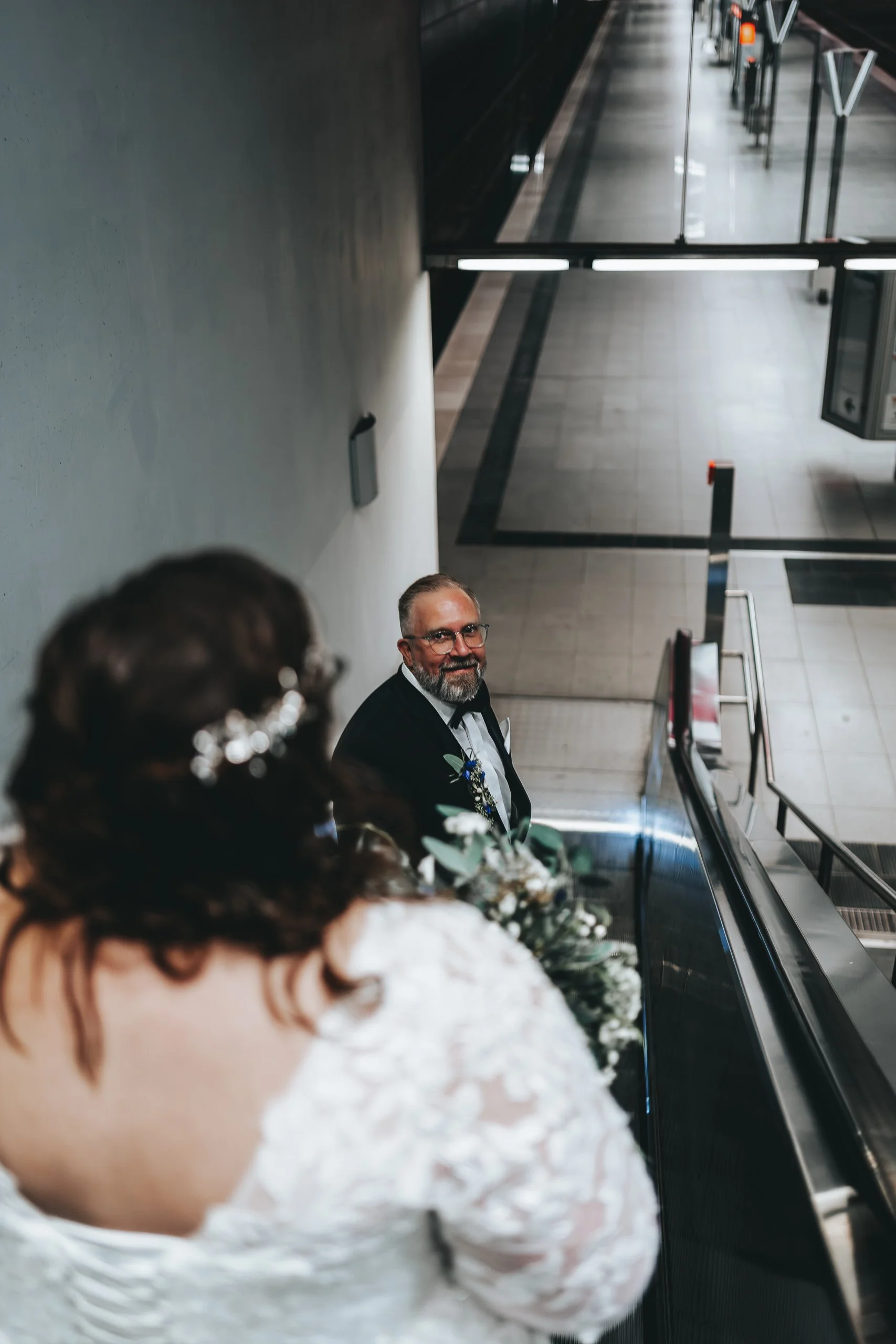 Braut mit dunklen Haaren und weißen Blumen in der Hand, ein lächelnder Mann in Anzug ist auf einer Rolltreppe im Hintergrund in einem modernen Gebäude sichtbar.