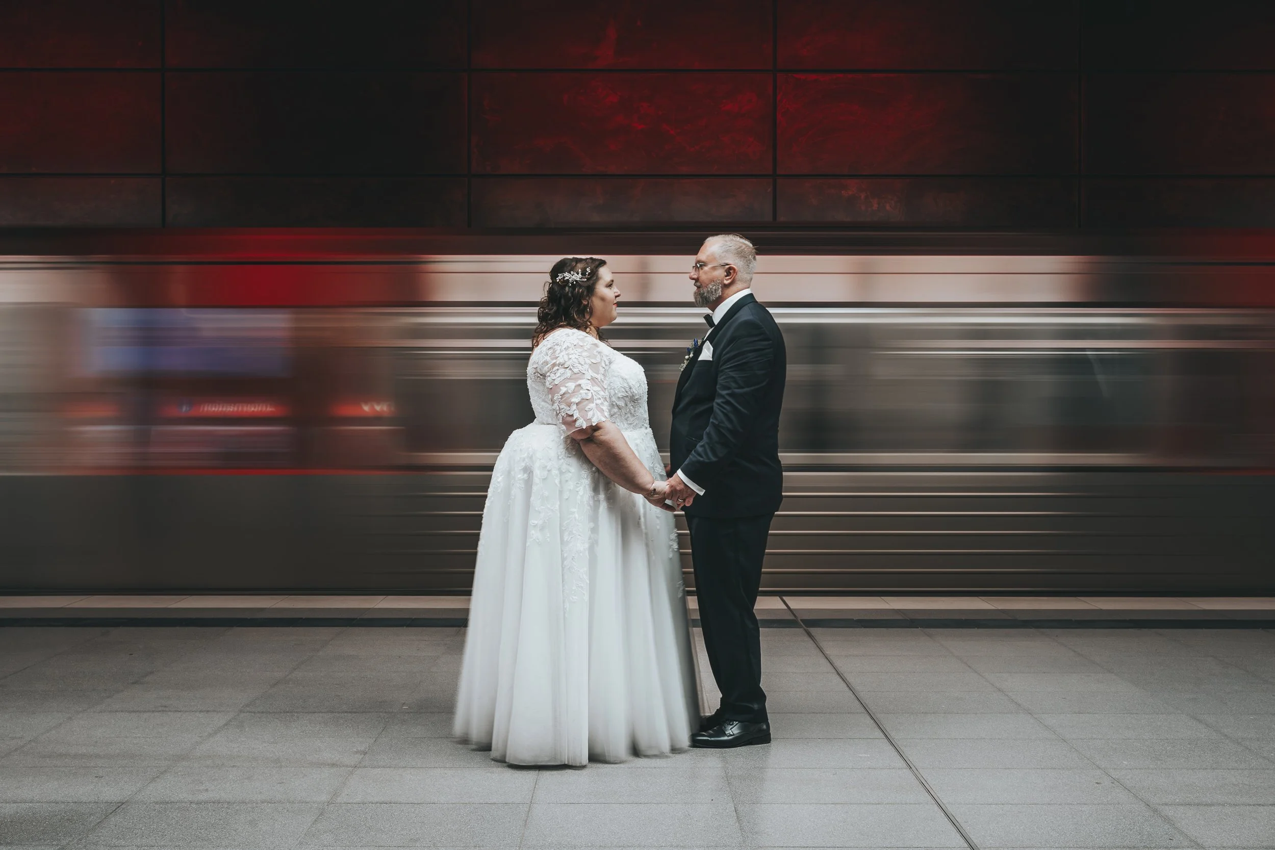 Ein Brautpaar in formeller Kleidung steht sich gegenüber vor einem laufenden Zug in einem unterirdischen Bahnhof, sie hält Hände und schaut sich an.