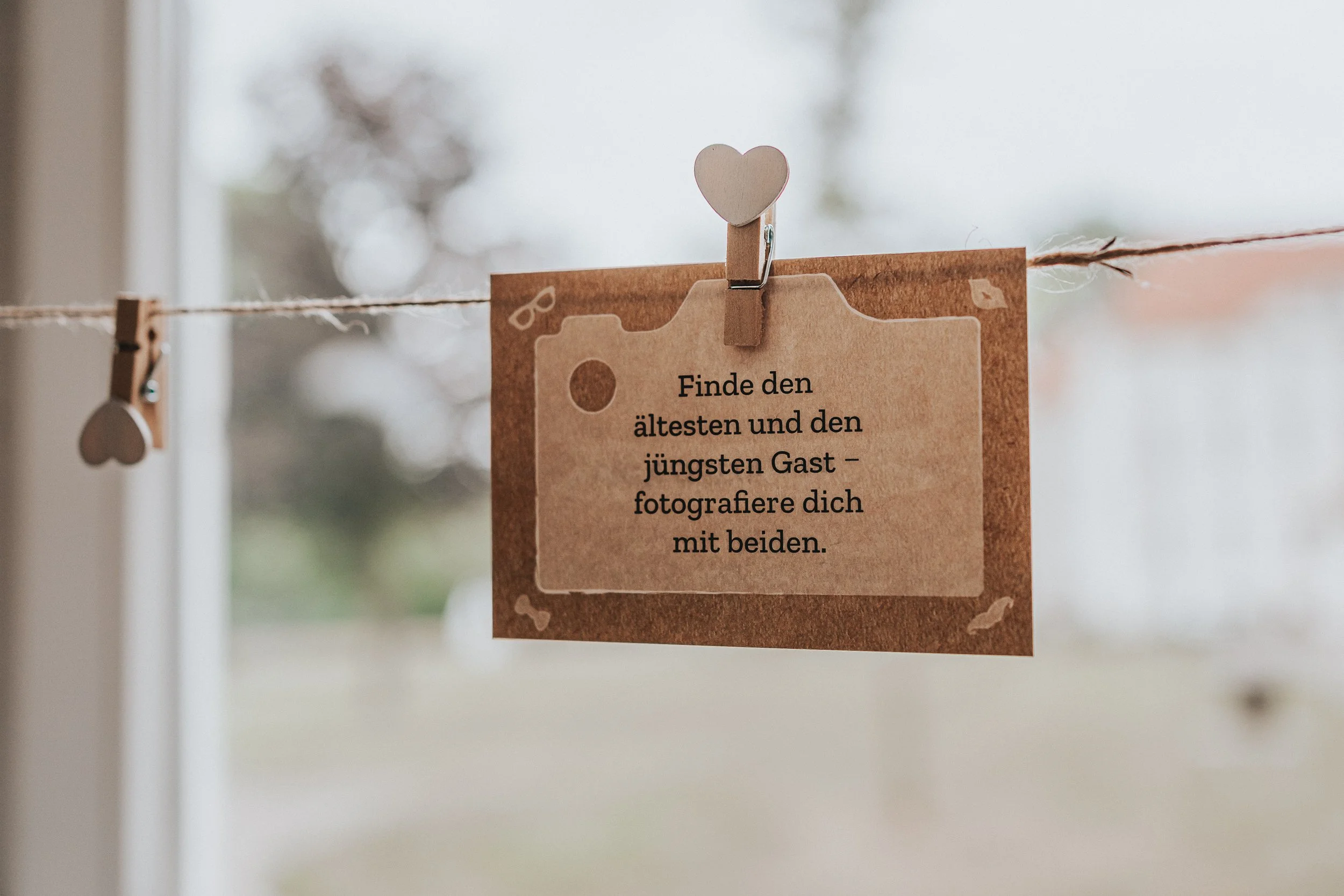 Ein Zettel hängt an einer Schnur, mit einem kleinen Holzherz am Clip. Der Zettel trägt eine Nachricht auf Deutsch, die besagt: 'Finde den ältesten und den jüngsten Gast – fotografiere dich mit beiden.'