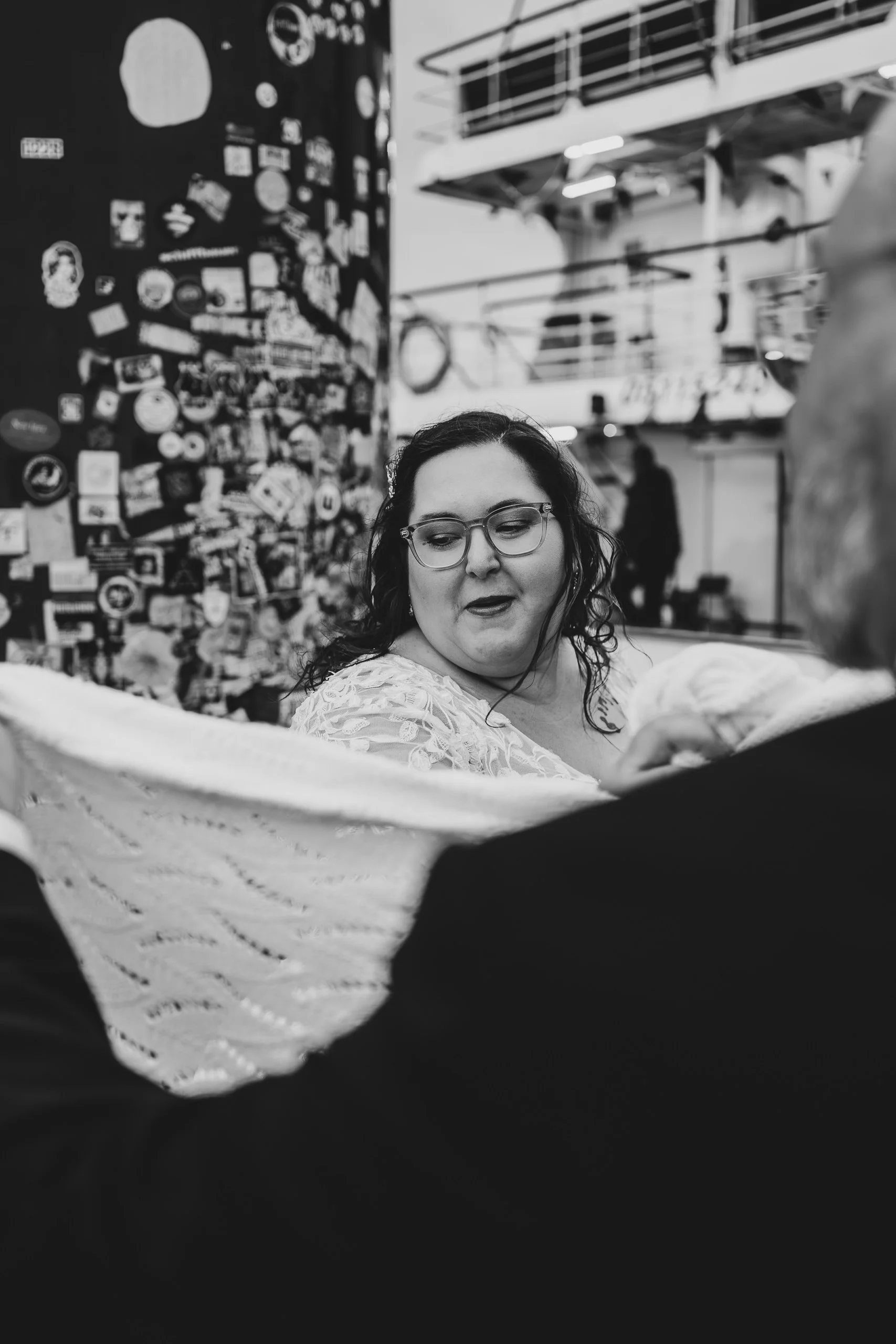 Schwarz-weiß-Fotografie einer Frau mit Brille, die einen Baby tragenden Arm betrachtet, in einer urbanen Umgebung mit einem Wand voller Aufkleber im Hintergrund.