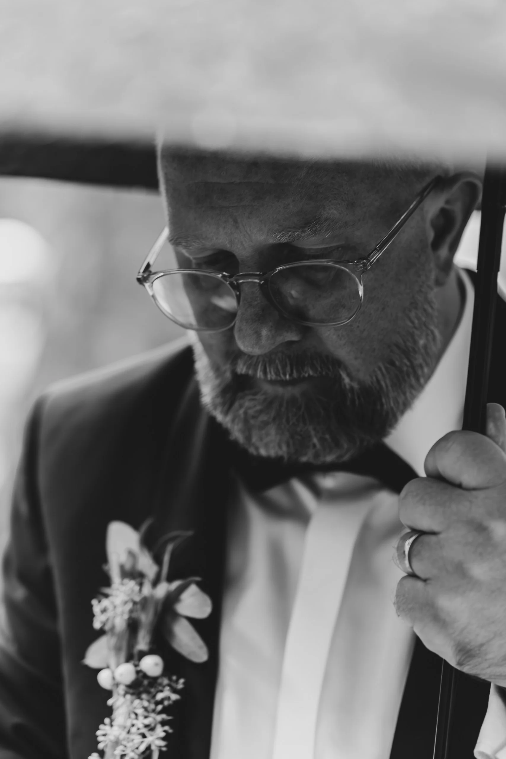 Schwarz-weiß-Foto eines Mannes mit Brille, formeller Kleidung und einer Schleife, der eine Blume trägt, und eine Hand hält einen Regenschirm über seinem Kopf.