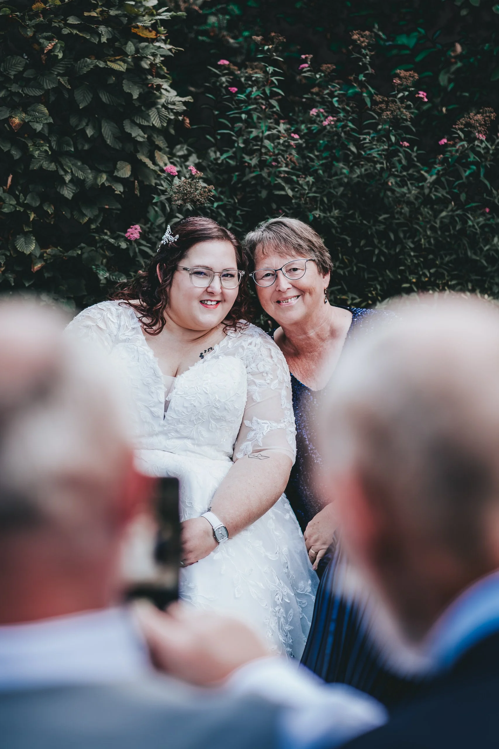 Zwei Frauen, eine junge in einem weißen Brautkleid und eine ältere in einem dunkelblauen Kleid, posieren lachend vor einem Busch mit pinken Blüten bei einer Hochzeit.