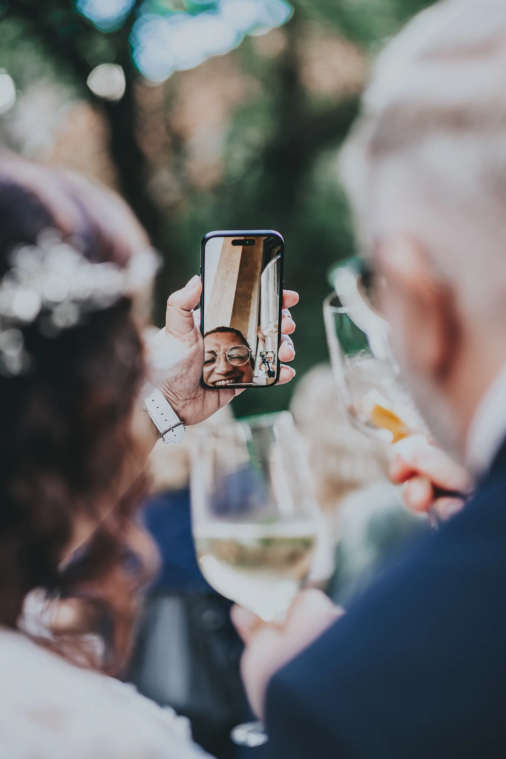 Menschen bei einer Feier, die mit einem Smartphone ein Foto machen, während sie Wein trinken. Der Fokus liegt auf dem Handy, das ein fröhliches Gesicht zeigt, während es ein Bild in einer lebhaften, natürlichen Umgebung aufnimmt.