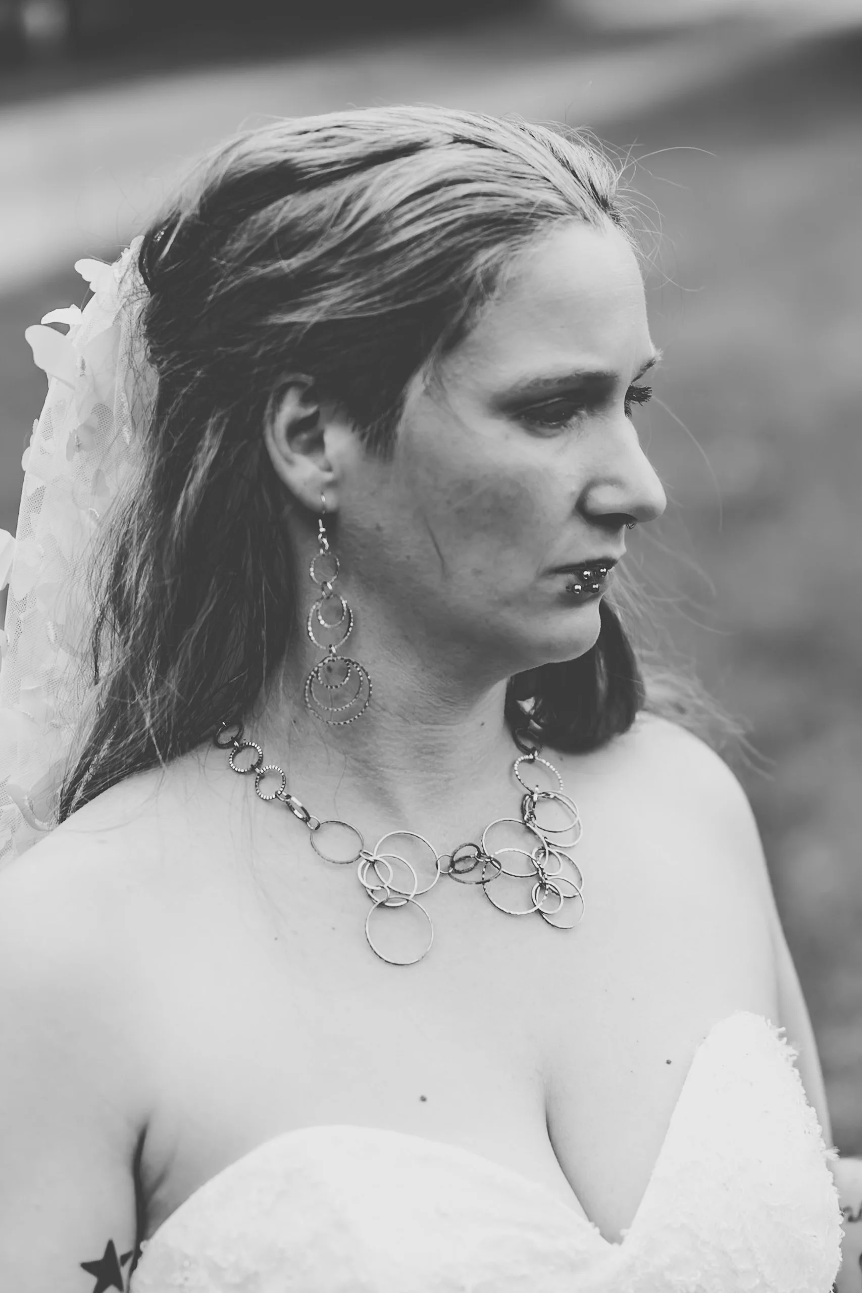 Schwarz-weiß-Foto einer Frau mit langen Haaren, die einen Hochzeitskleid trägt, traditionellen Schmuck wie Ohrringe und Halskette; sie hat einen Schleier im Haar. Der Hintergrund ist unscharf.