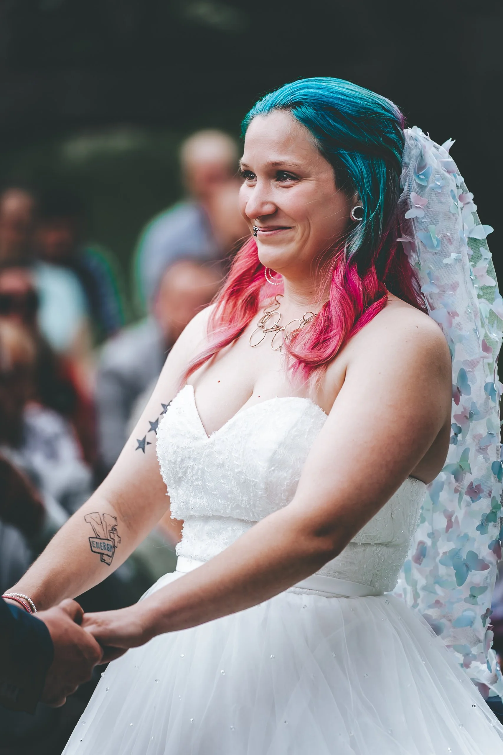 Eine Frau in Hochzeitskleid mit buntem, zottelig gefärbtem Haar beim Heiratsantrag, die jemandem ihre Hände hält, im Hintergrund eine Gruppe von Menschen, draußen im Grünen.