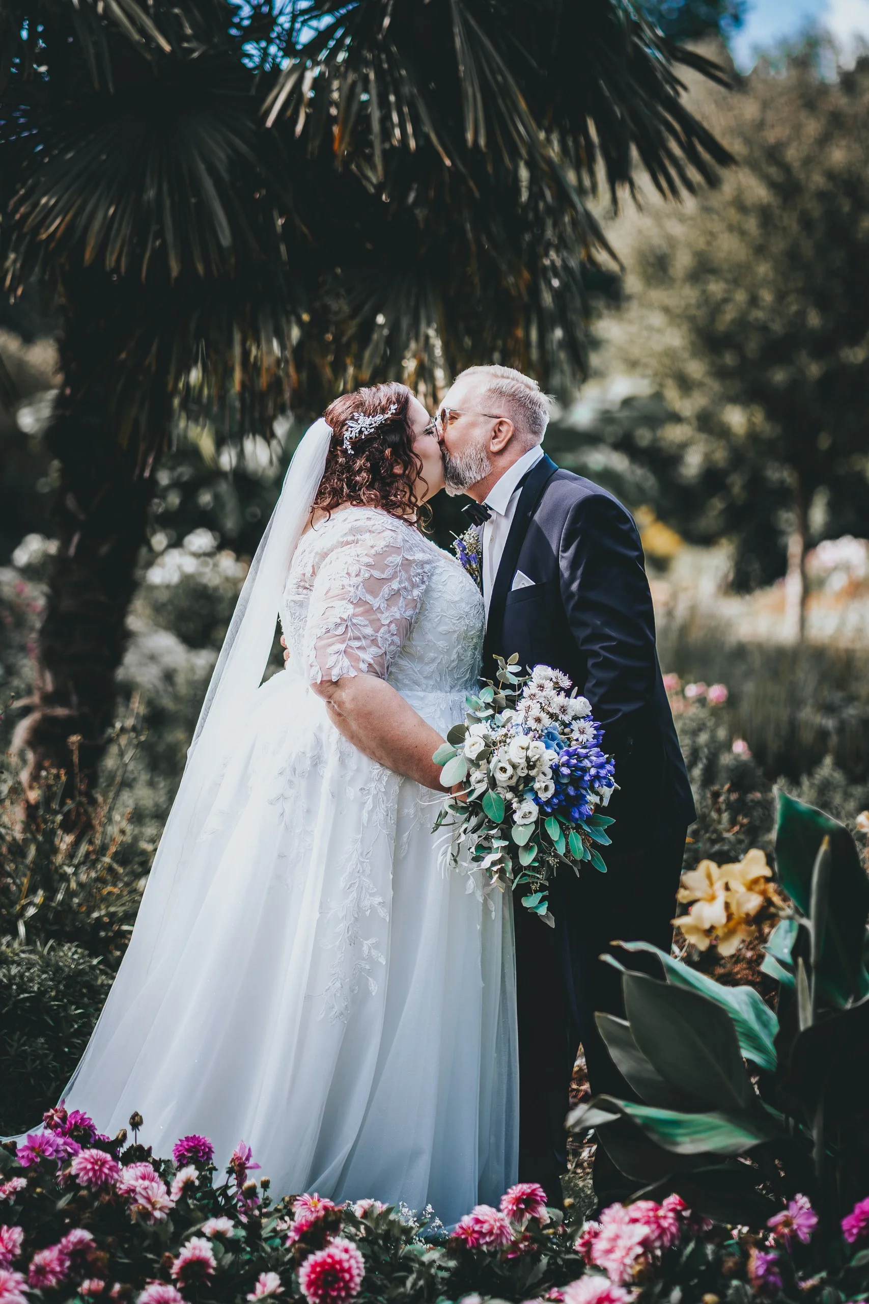 Ein Brautpaar bei einer Hochzeit im Grünen, sie küssen sich unter einem Baum, umgeben von Blumen, trägt elegante Kleidung, die Frau trägt ein weißes Brautkleid mit Schleier, der Mann trägt einen dunklen Anzug mit Fliege.