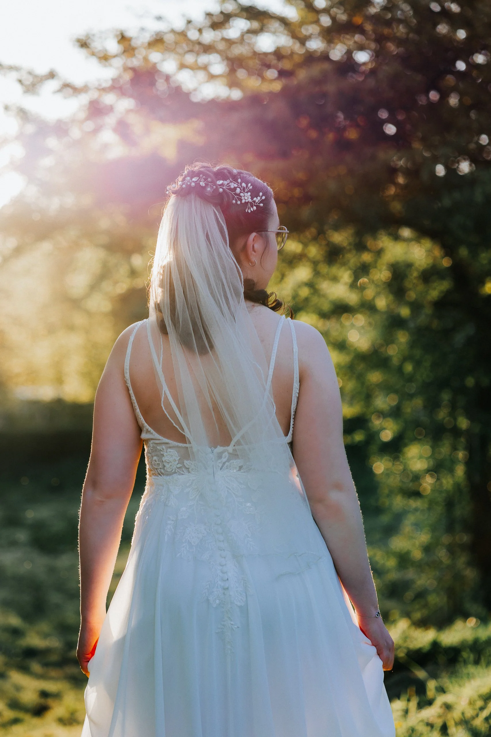 Eine Frau in einem weißen Brautkleid mit Schleier und Blumenkranz steht im Sonnenlicht in einem Garten, von hinten fotografiert.