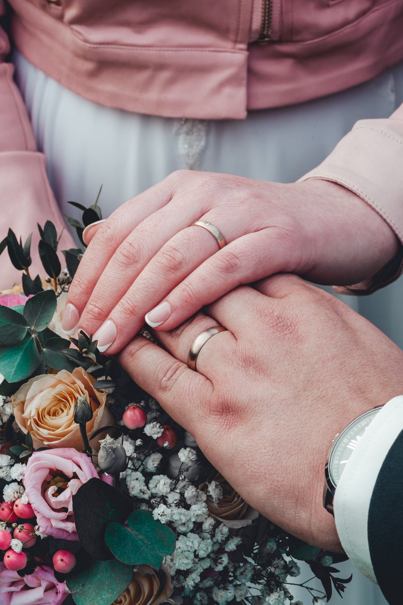 Zwei Menschen halten sich bei einer Hochzeit oder Feier die Hände, beide tragen Eheringe, im Hintergrund bunte Blumen, darunter Rosen, Beeren und lokale Blüten.