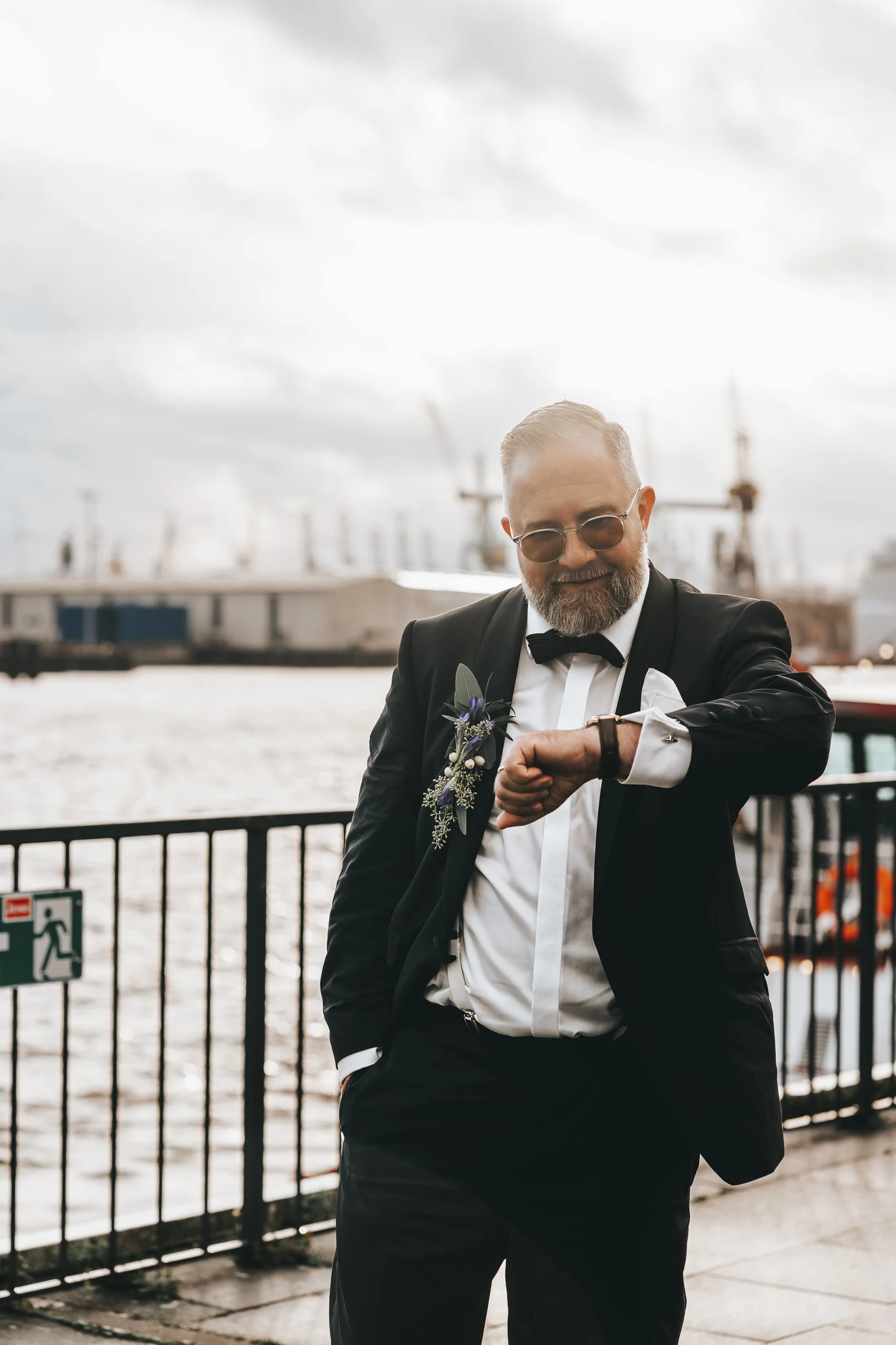 Bräutigam in formeller Kleidung mit Brille und Fliege schaut beim After-Wedding-Shooting im Hamburger Hafen auf seine Armbanduhr, Schiffe und Wasser im Hintergrund.
