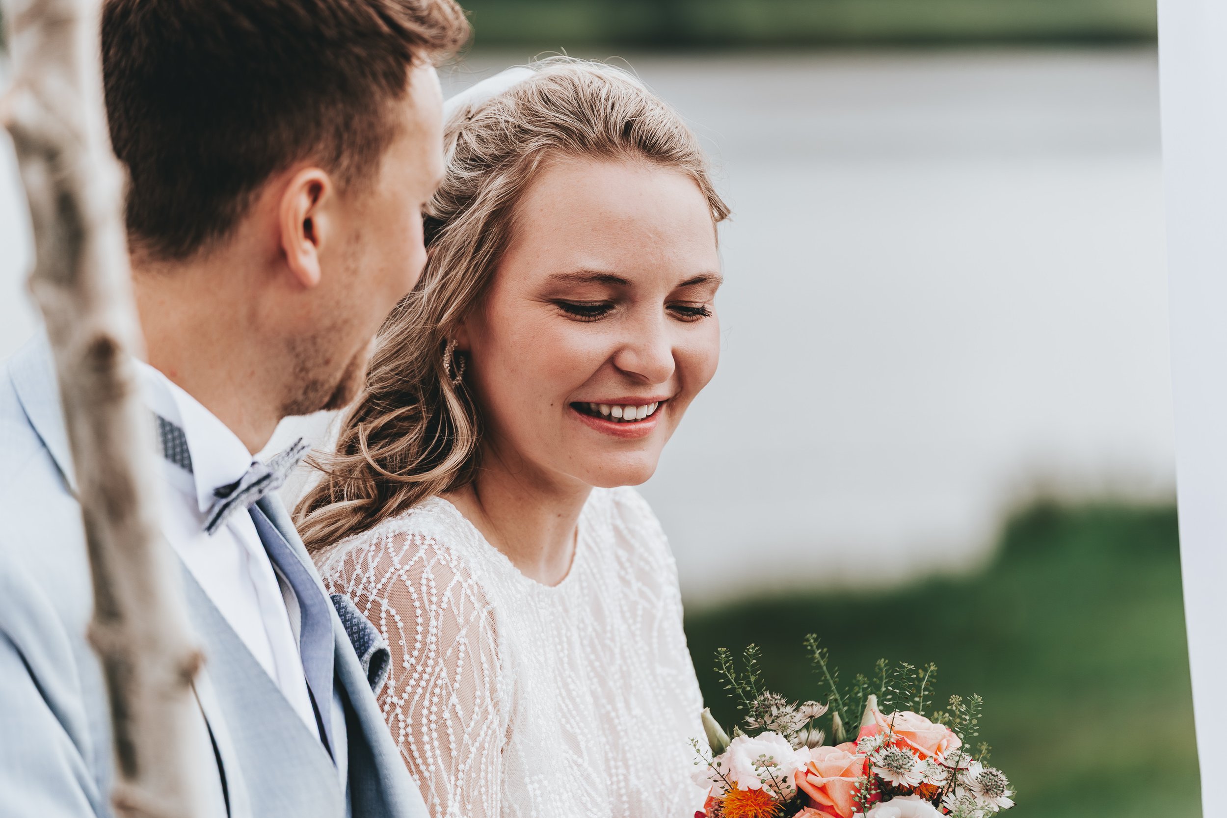 Ein Brautpaar bei einer Hochzeit im Freien, lächelnd, mit Blumen, im Hintergrund Wasser und Natur.