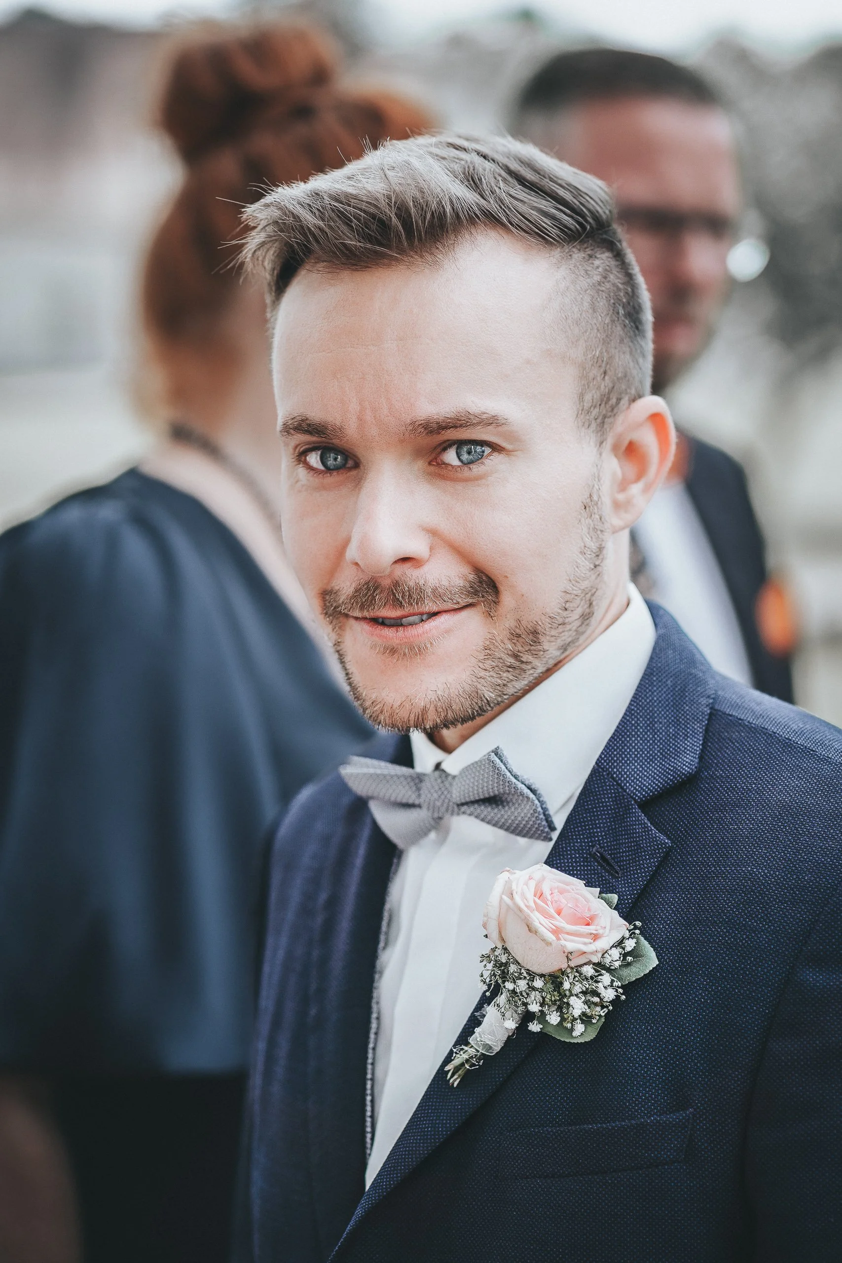 Bräutigam in Hochzeitsanzug mit Fliege und boutonniere, blickt in die Kamera, im Hintergrund andere Personen bei einer Hochzeit.