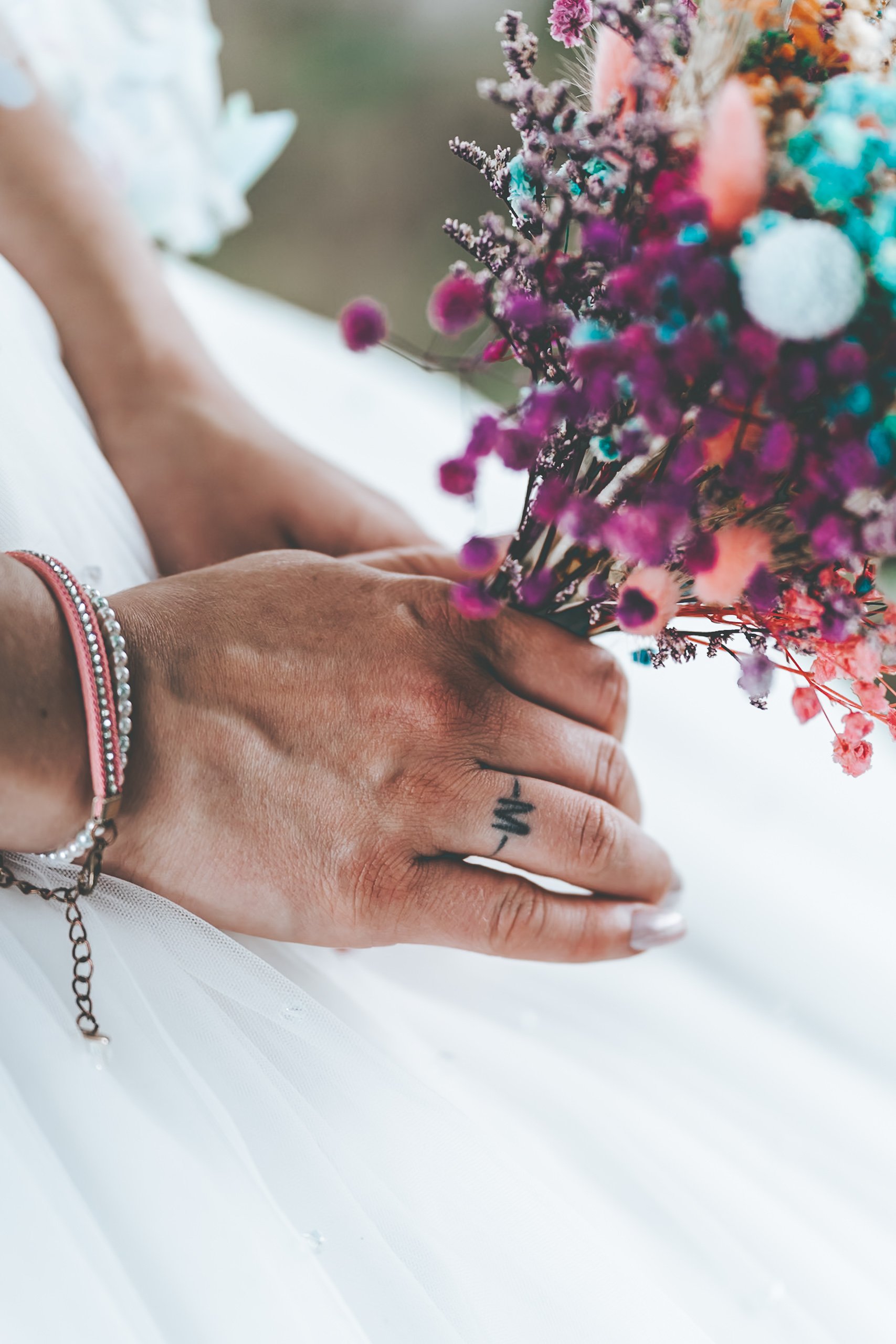 Hand halte einen Blumenstrauß, getragen mit mehreren Armbändern und einem kleinen Tattoo auf dem Finger, auf einem weißen Stoffhintergrund.