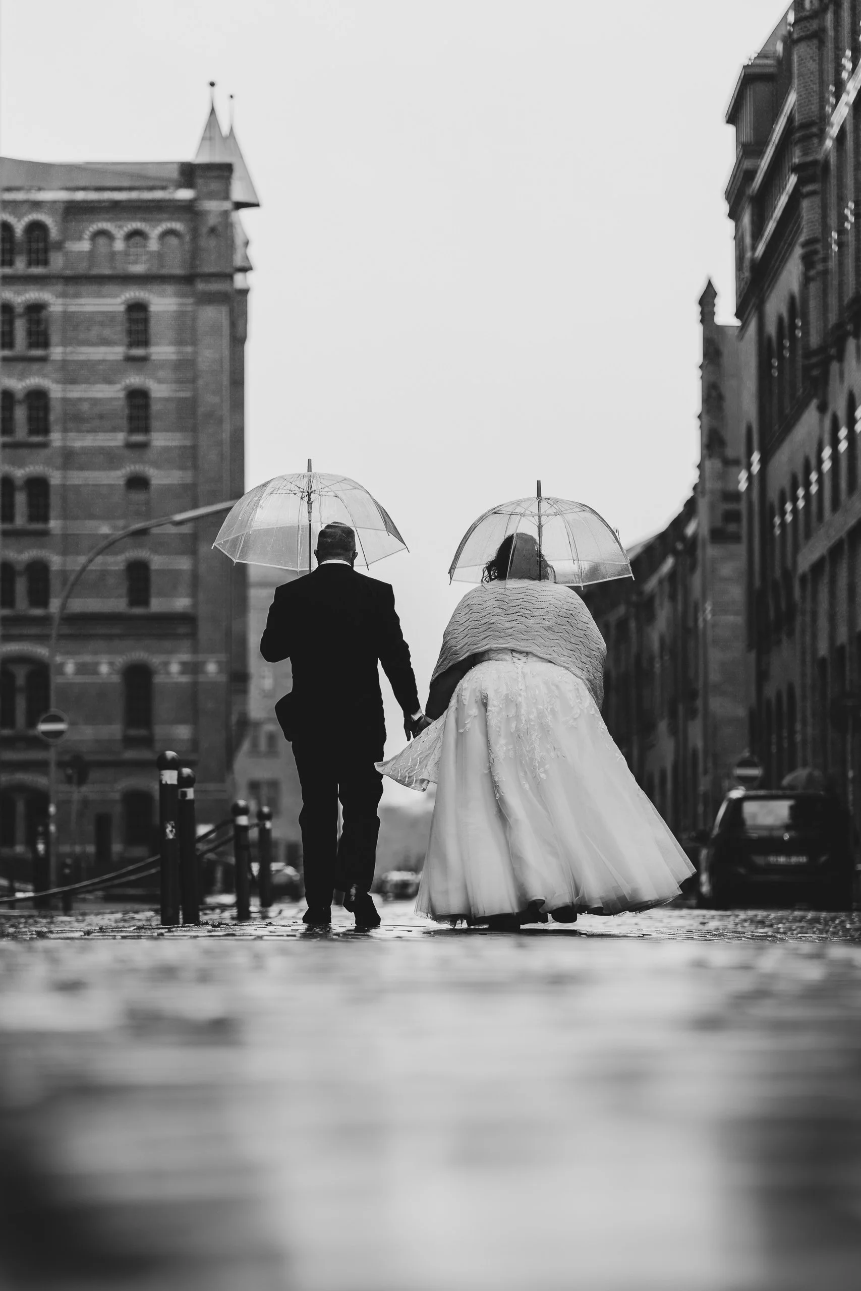 Ein Brautpaar läuft bei Regen durch eine Stadt, beide tragen Regenschirme, sie hält seine Hand, sie trägt ein langes Hochzeitskleid, er einen Anzug, die Umgebung ist städtisch mit alten Gebäuden.